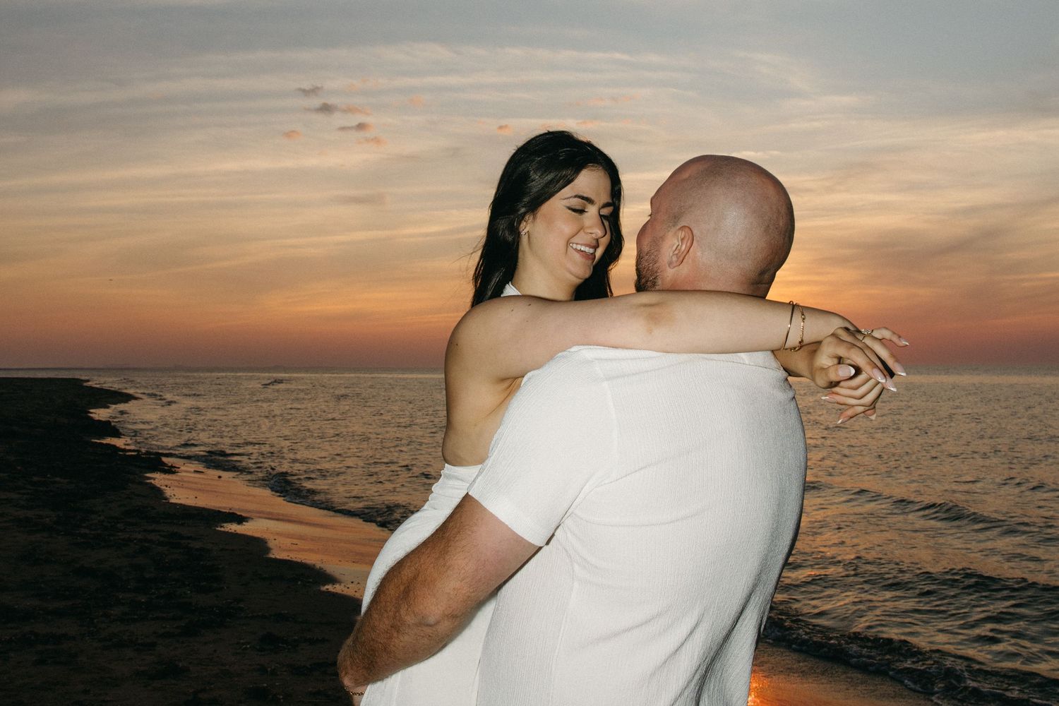Couple embracing on beach at sunset, woman in white dress laughing joyfully, golden sky over ocean backdrop.