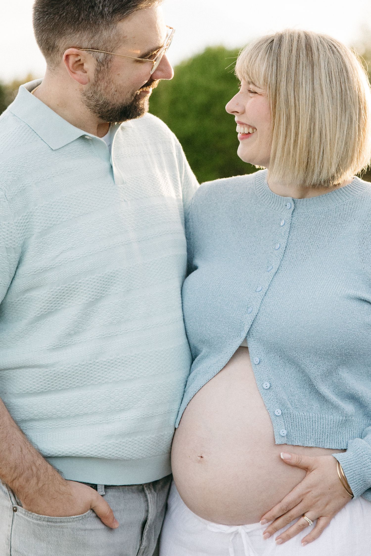 Happy pregnant couple in matching light blue outfits smiling at each other outdoors, woman cradling baby bump.