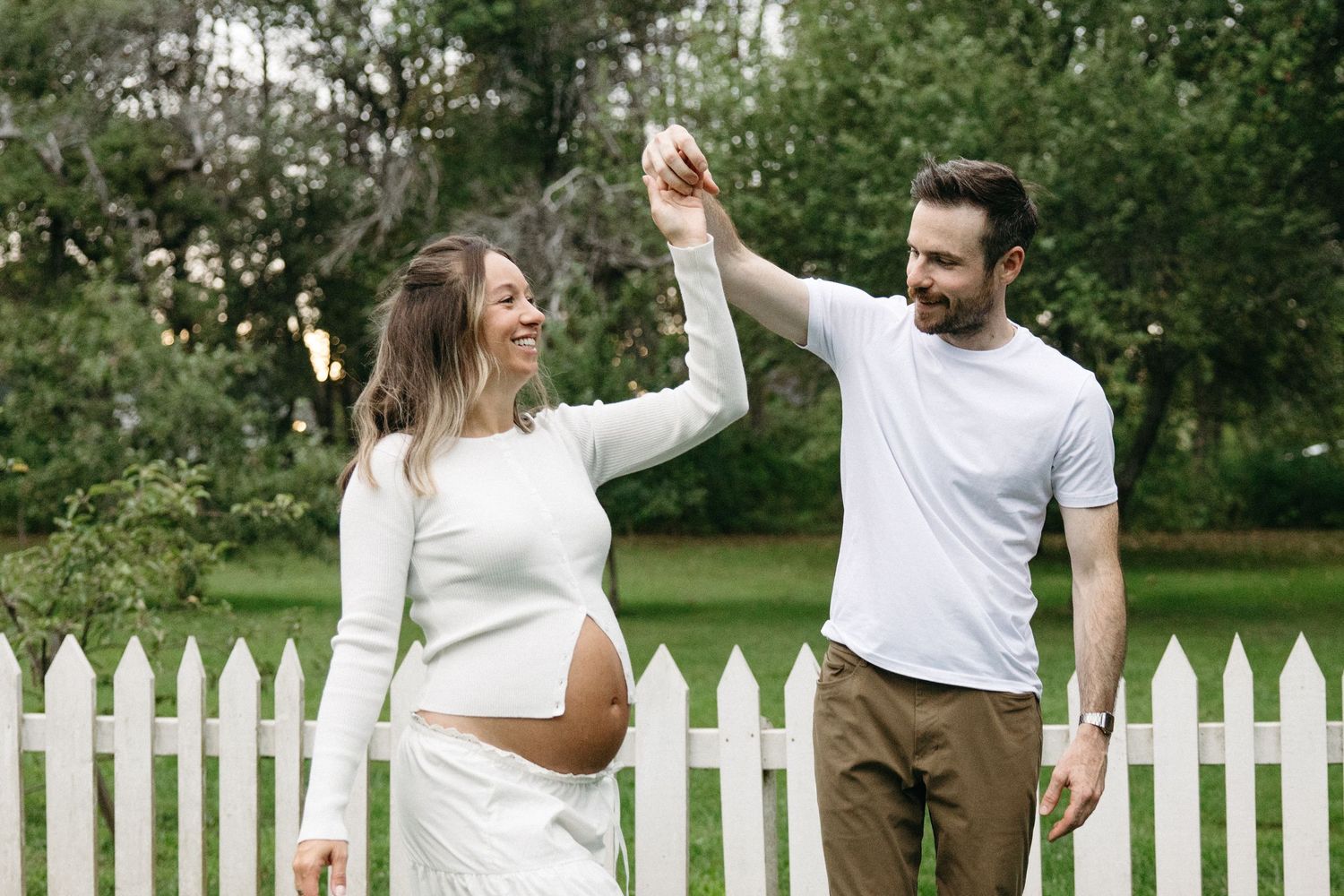 Smiling pregnant woman in white outfit dances with bearded man near white picket fence in green garden.