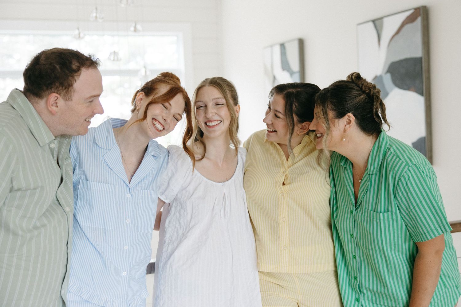 Five friends laughing and embracing indoors, wearing colorful pastel pajamas in blue, white, yellow, and green stripes.