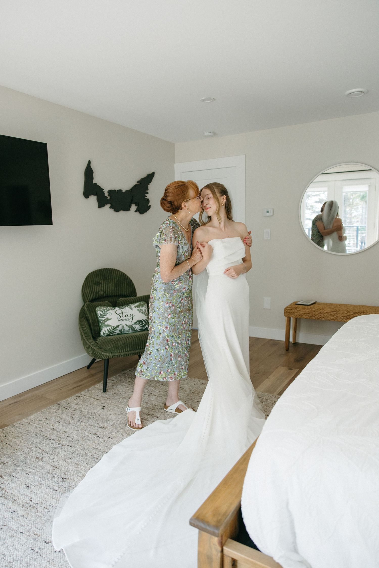 Mother kisses bride in elegant strapless wedding gown with long train in bright bedroom.