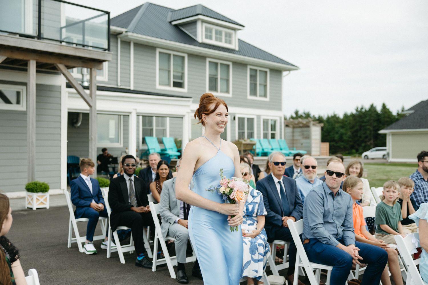 Bridesmaid in light blue dress walks down the aisle at an outdoor wedding ceremony with seated guests.