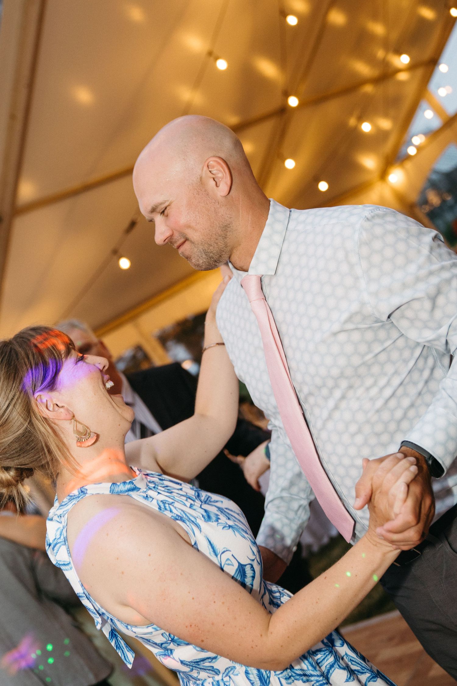 Bald groom dances with woman in floral dress under string lights at wedding reception.