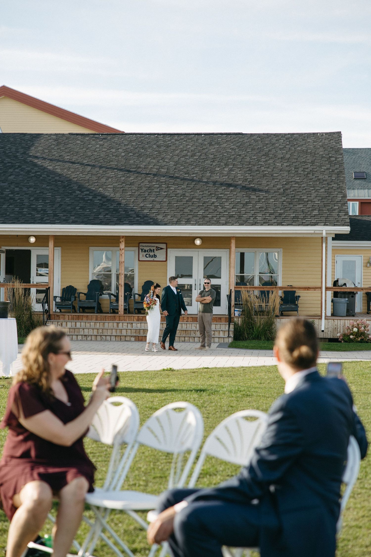 Wedding guests seated on white folding chairs watch an outdoor ceremony on the lawn in front of a yellow clubhouse with a covered porch.