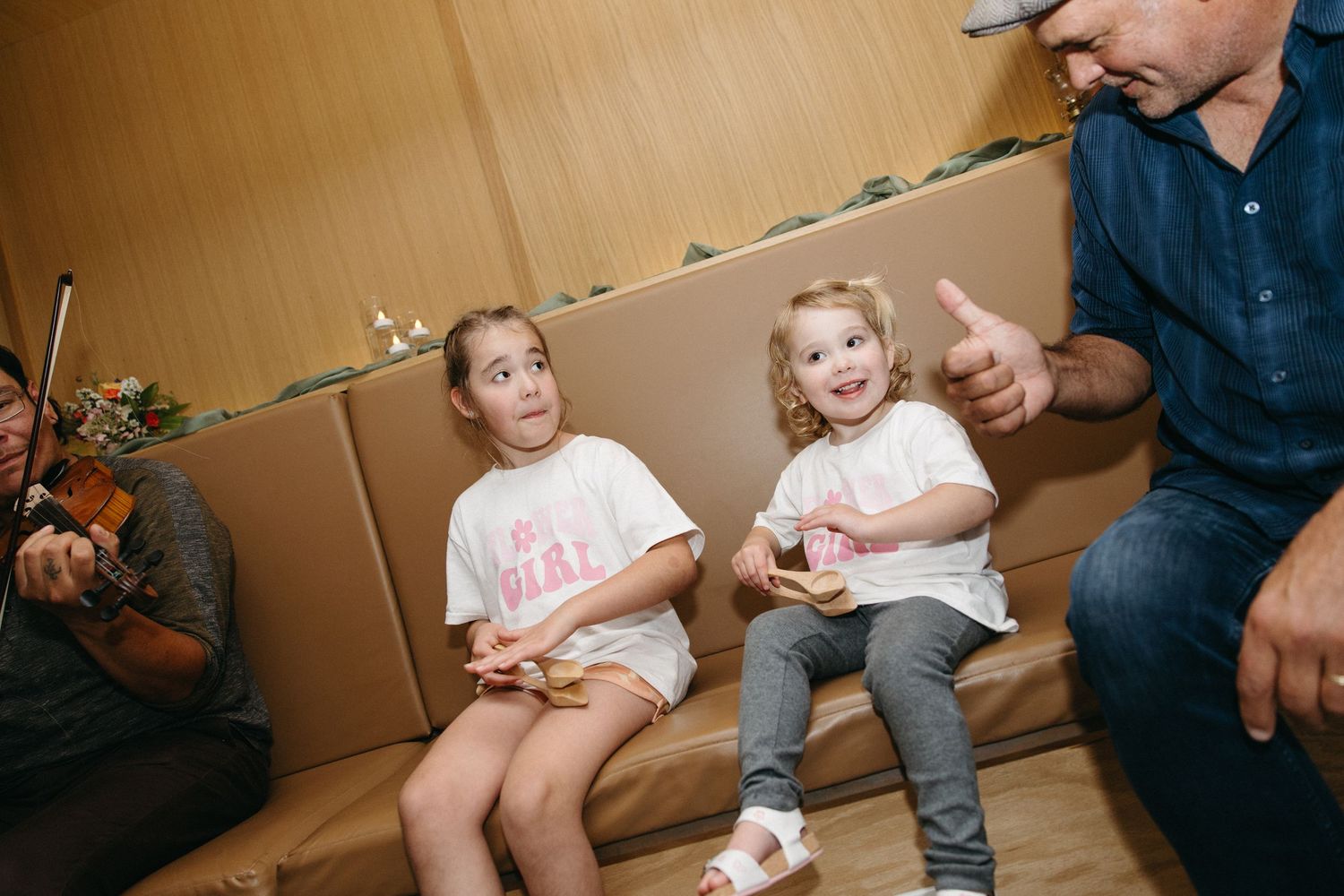 Two smiling young girls in white shirts sit on a tan booth bench while a man gives a thumbs up and a violinist plays beside them.