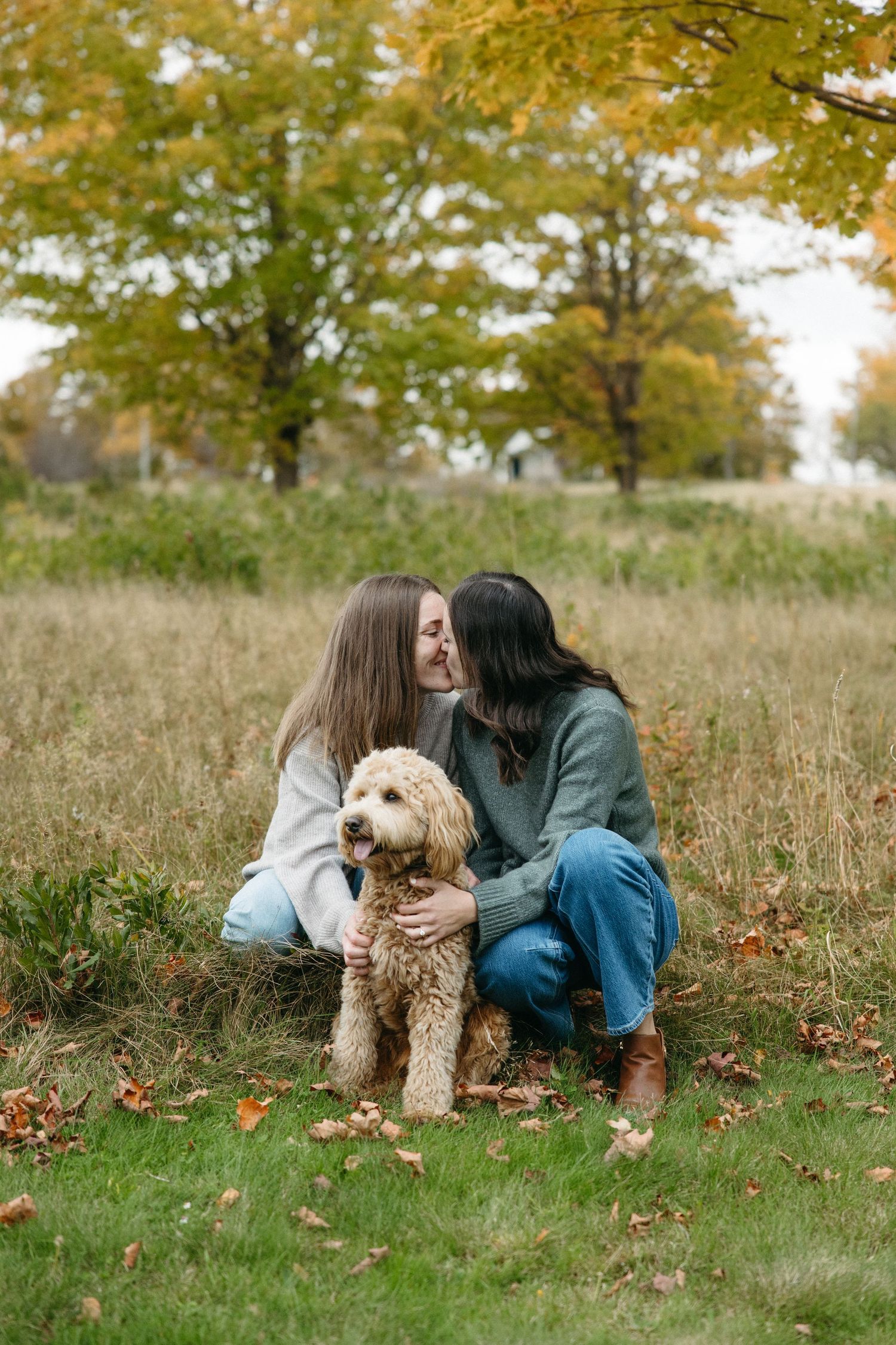 Two women kiss while kneeling in a grassy autumn field with a fluffy goldendoodle between them, surrounded by fall foliage.