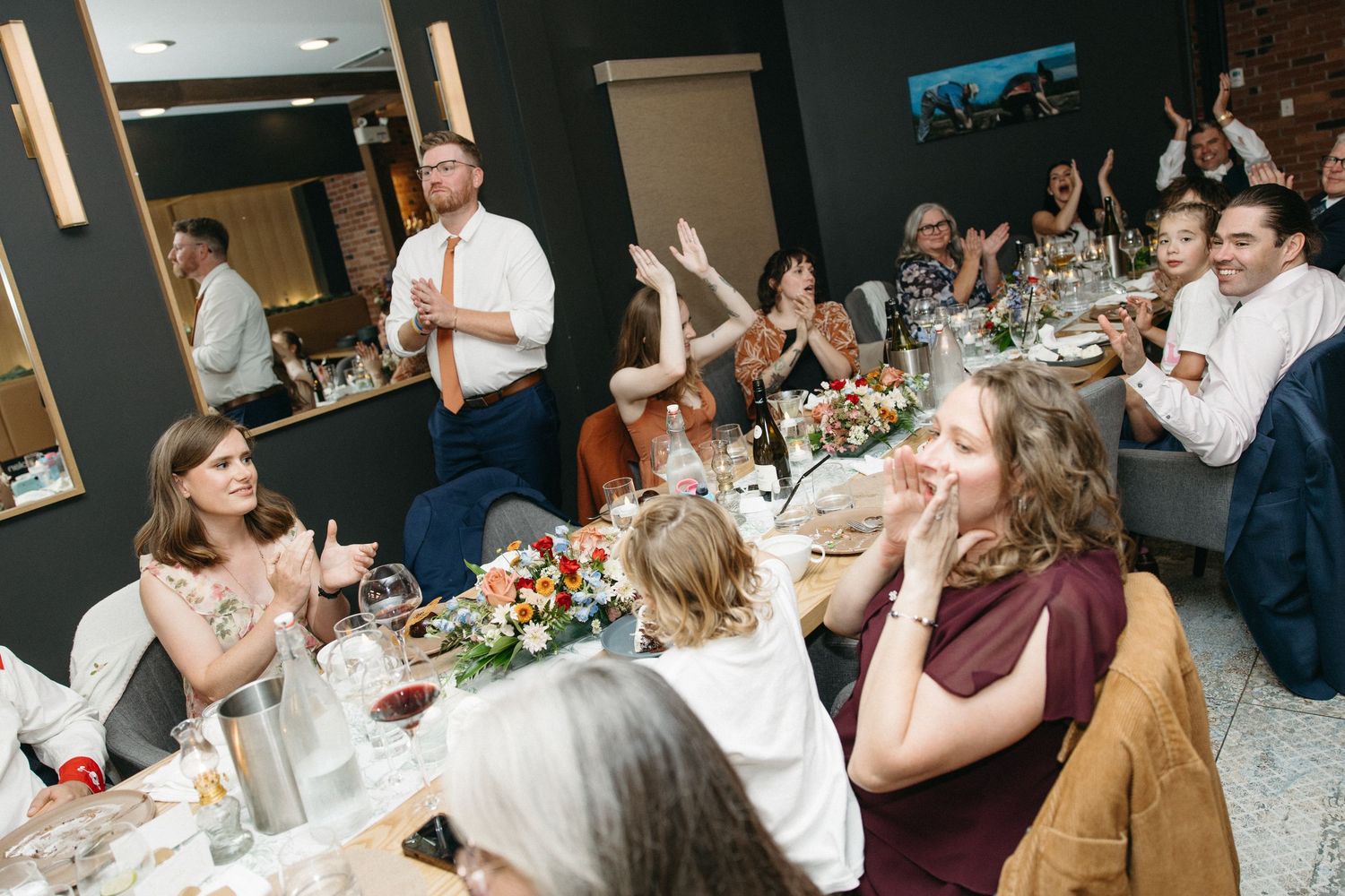 Wedding guests laugh and applaud around a decorated banquet table as a man in a tie gives a toast at a lively reception.