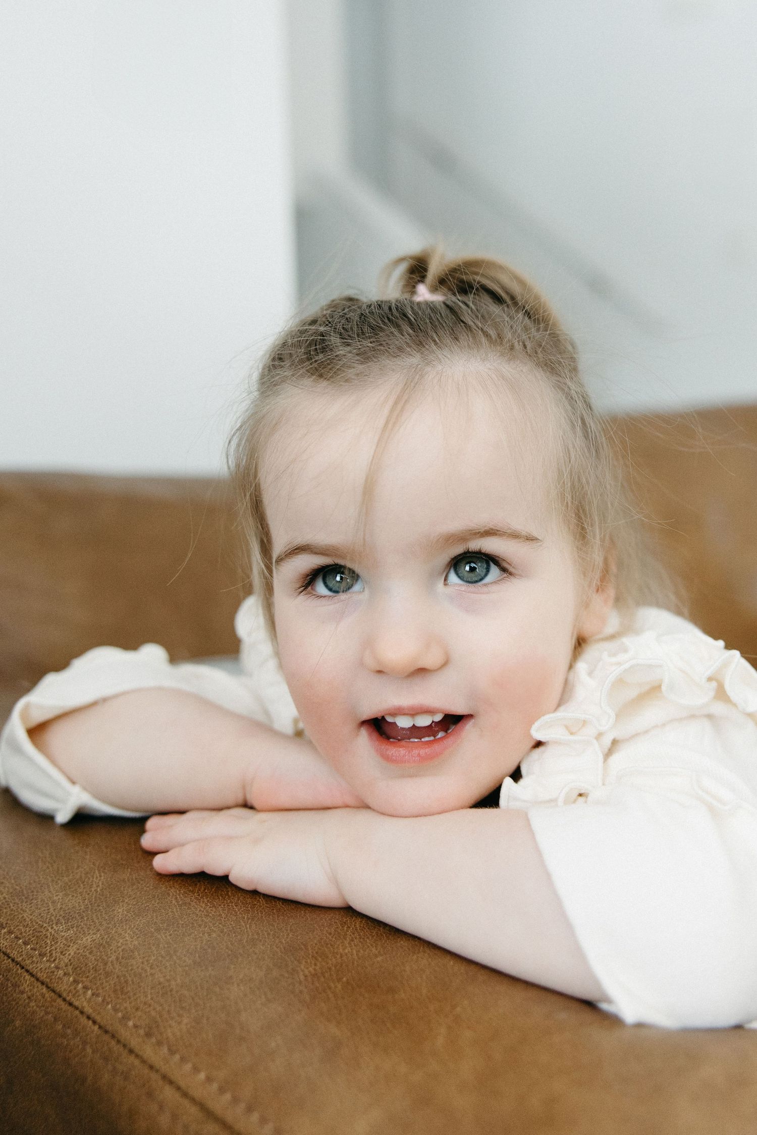 A young child in white peeks playfully over a brown couch cushion with bright eyes and a smile.