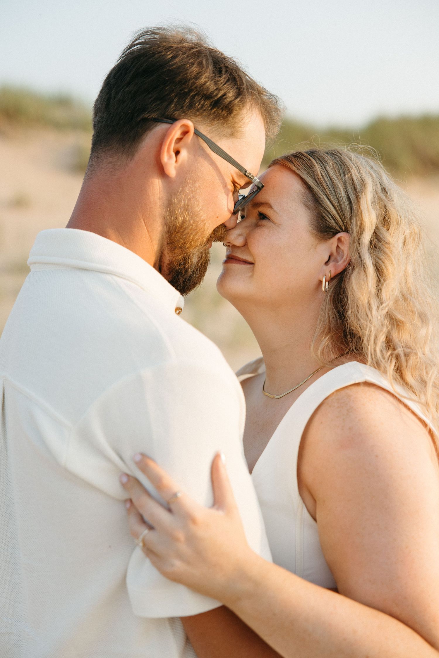 Romantic couple embracing on a sunny beach, woman smiling up at bearded man wearing sunglasses in white outfits.
