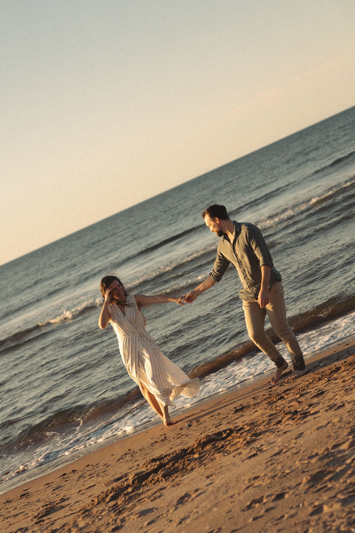 A couple playfully dances and holds hands on a sandy beach at golden sunset with ocean waves behind them.