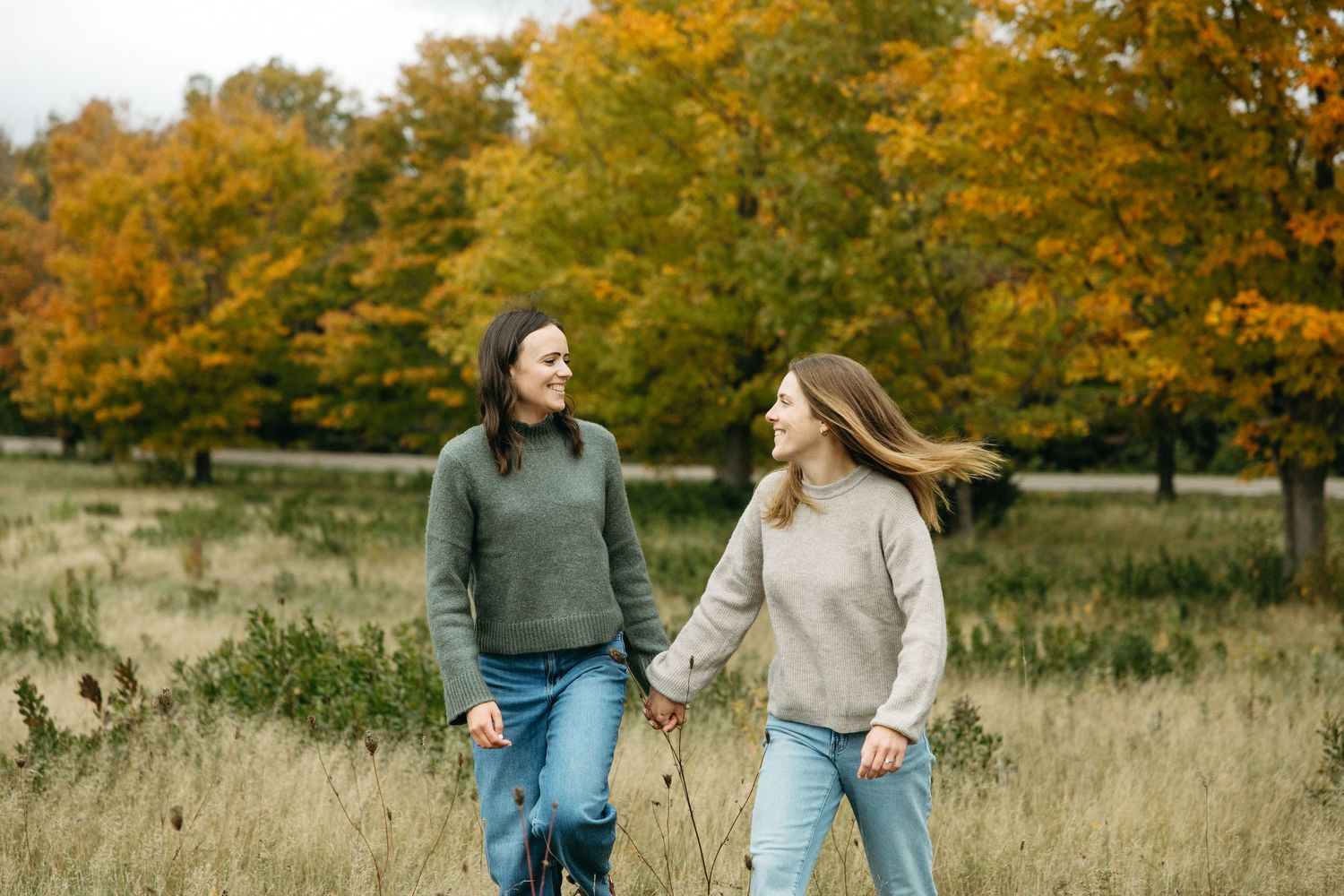 Two women holding hands and smiling while walking through a grassy field with colorful autumn trees in the background.