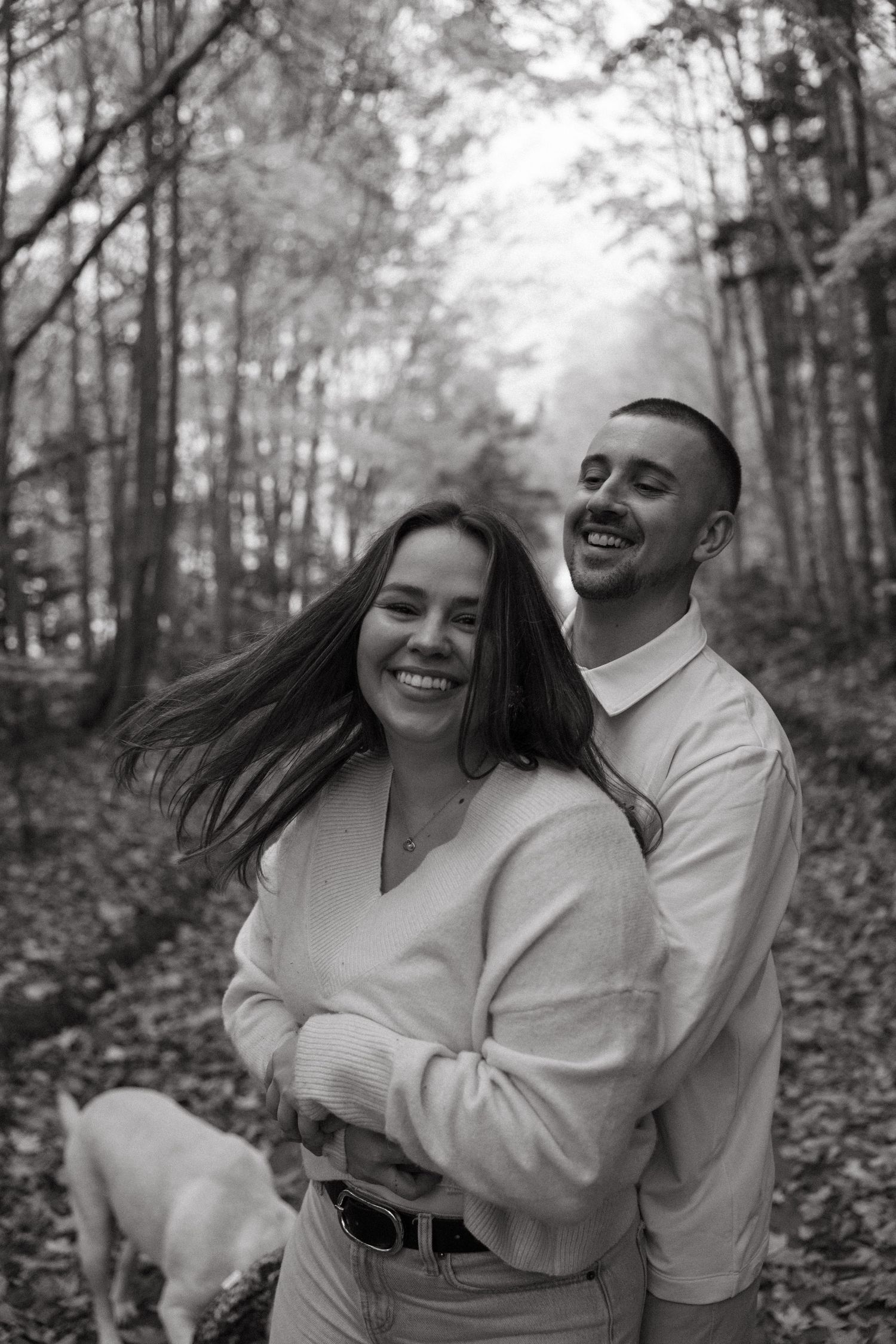 A couple shares joyful laughter while walking through a wooded path covered in autumn leaves.