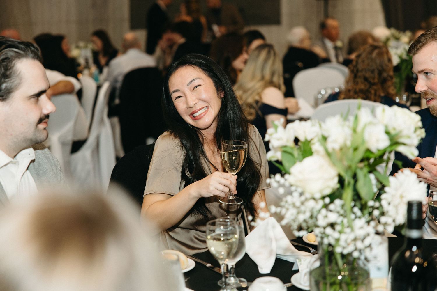 Wedding guests enjoying dinner conversation at an elegant reception with white floral centerpieces.