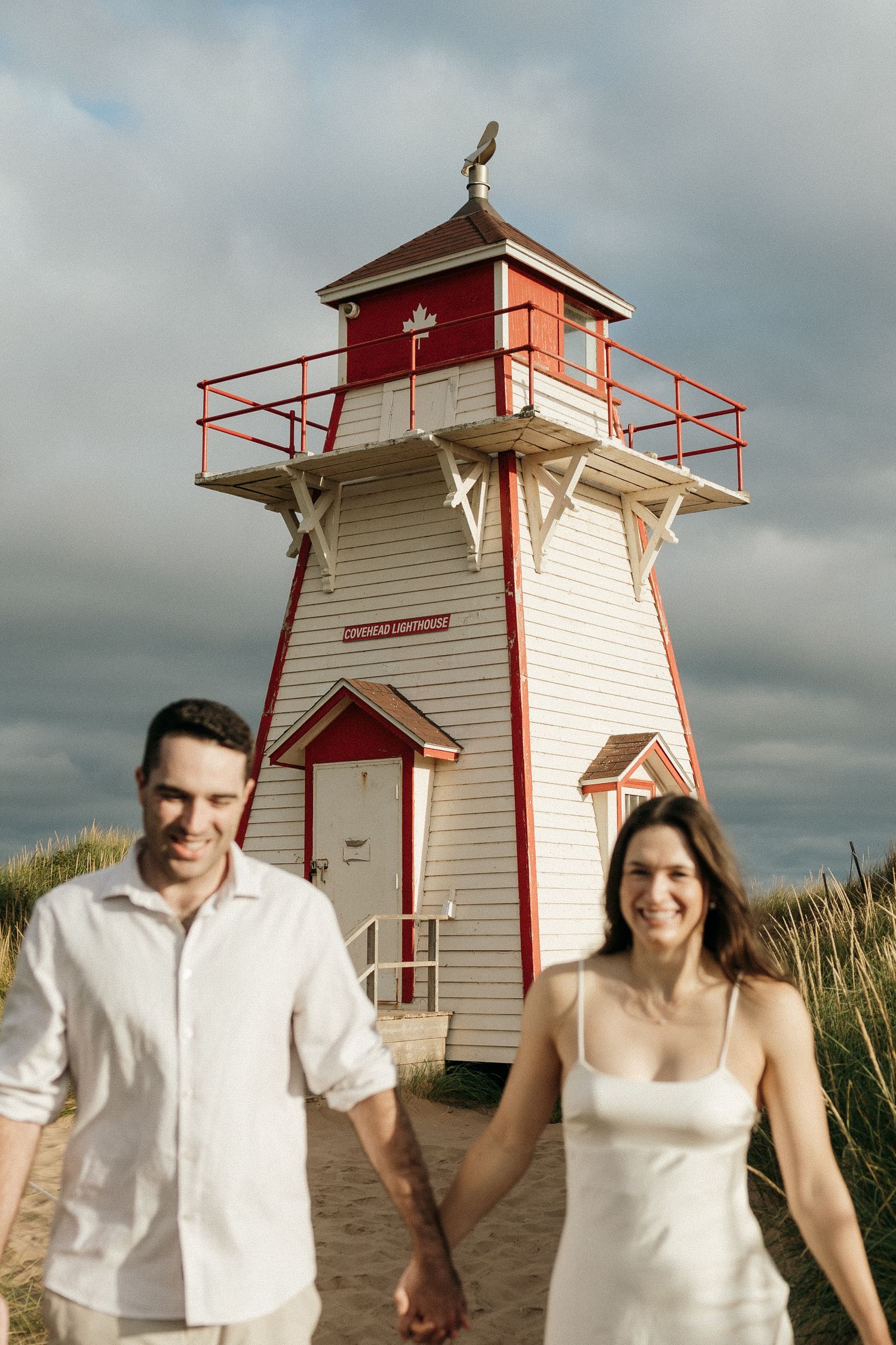 Couple in white clothing poses near red and white lighthouse on sandy beach.