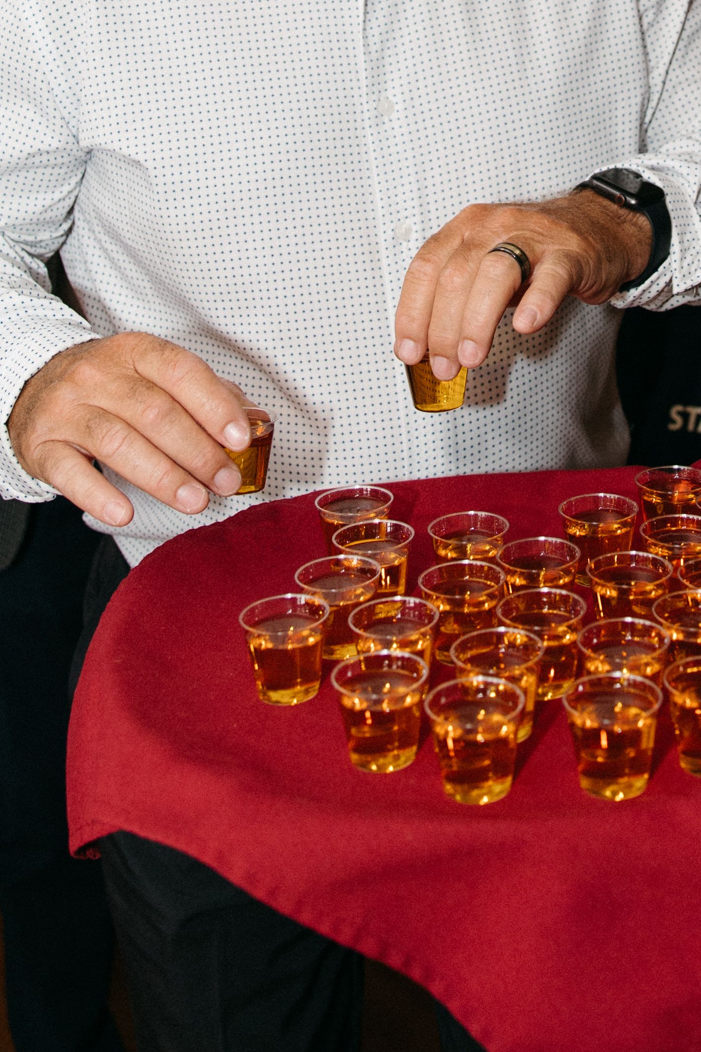 Man arranging rows of amber whiskey shots in plastic cups on a red velvet table at a wedding reception.