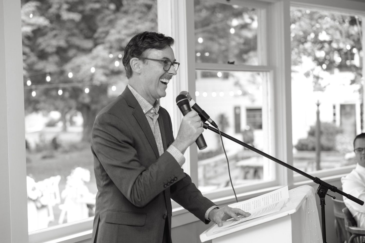 Man in suit delivers speech at microphone during wedding reception in black and white photo.