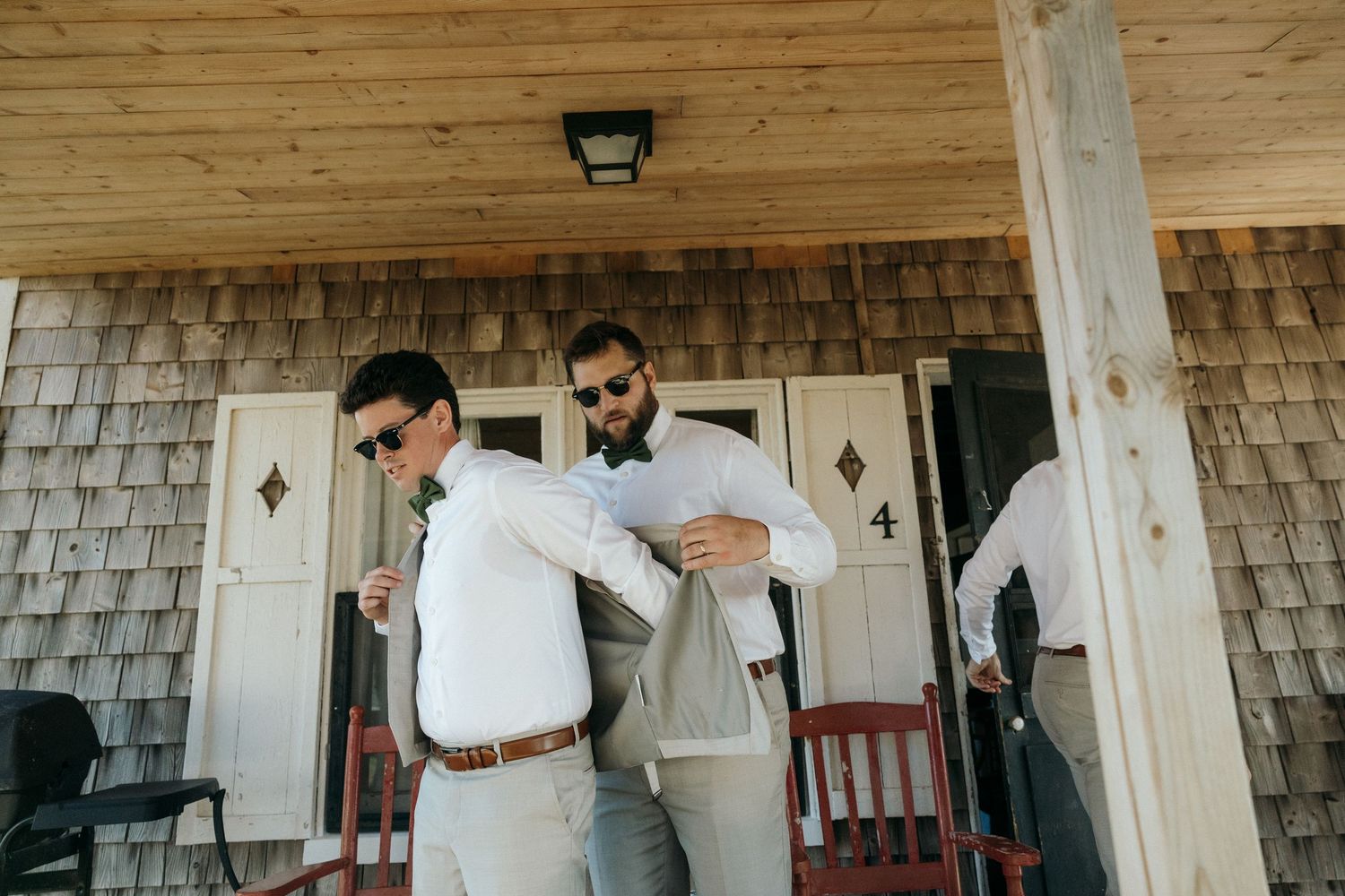 Two groomsmen in gray suits and sunglasses help each other get dressed on a rustic wooden cabin porch.