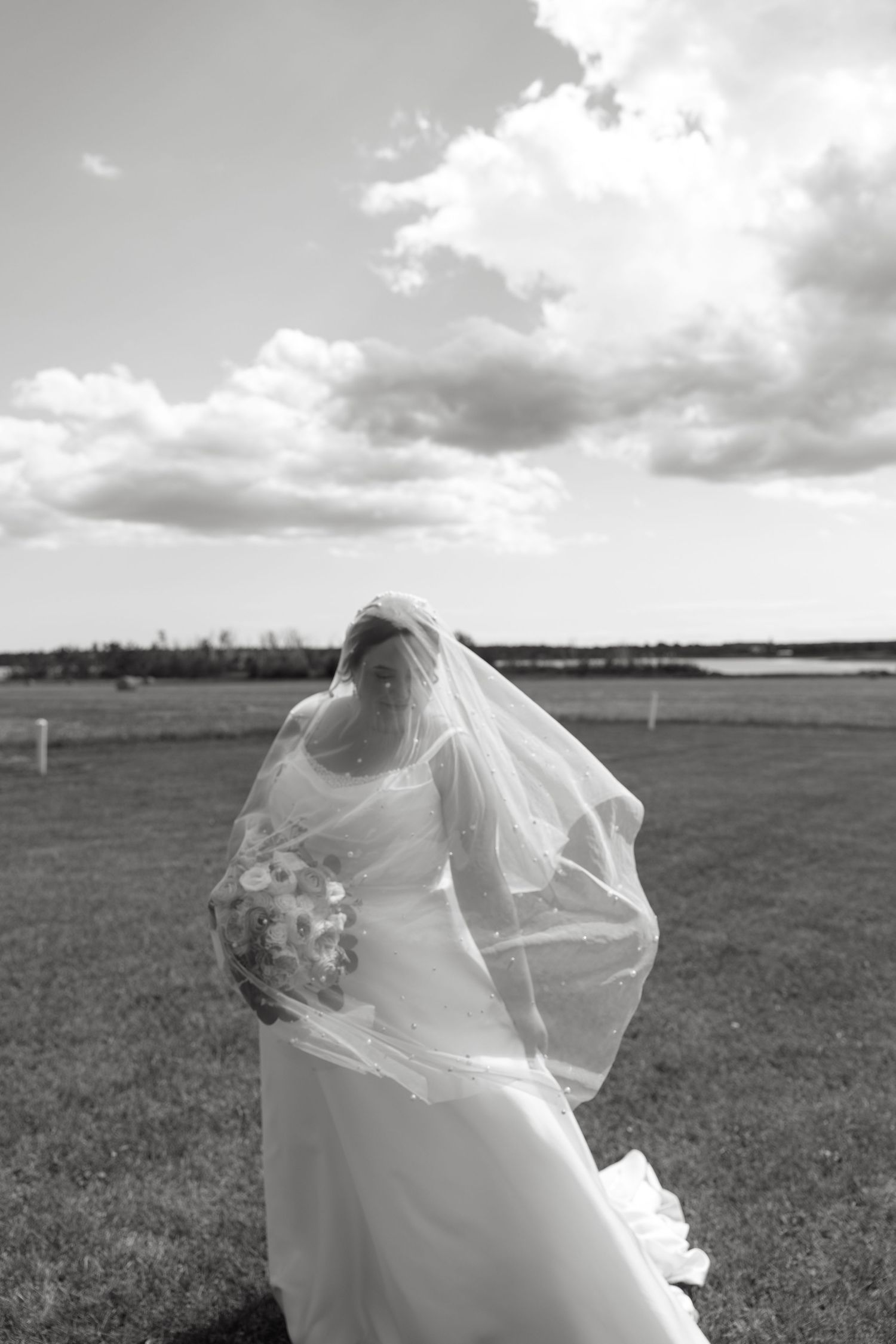 Bride in white wedding gown and flowing veil holds bouquet in black and white outdoor field photo.