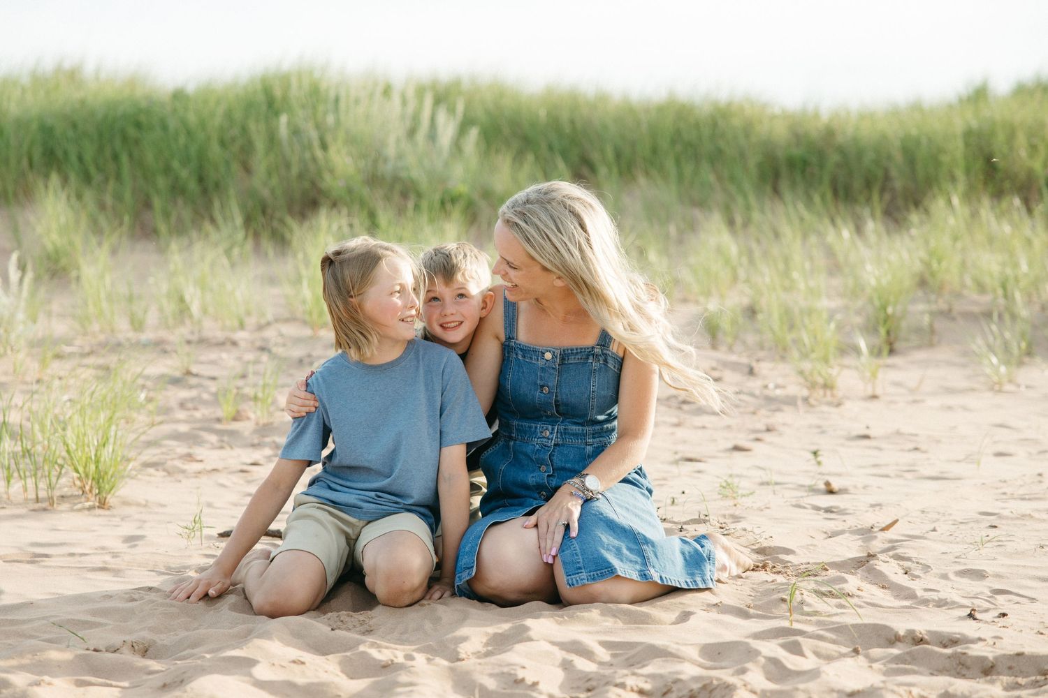 A blonde mother in a denim dress sits on sand smiling at her two children on a beach.