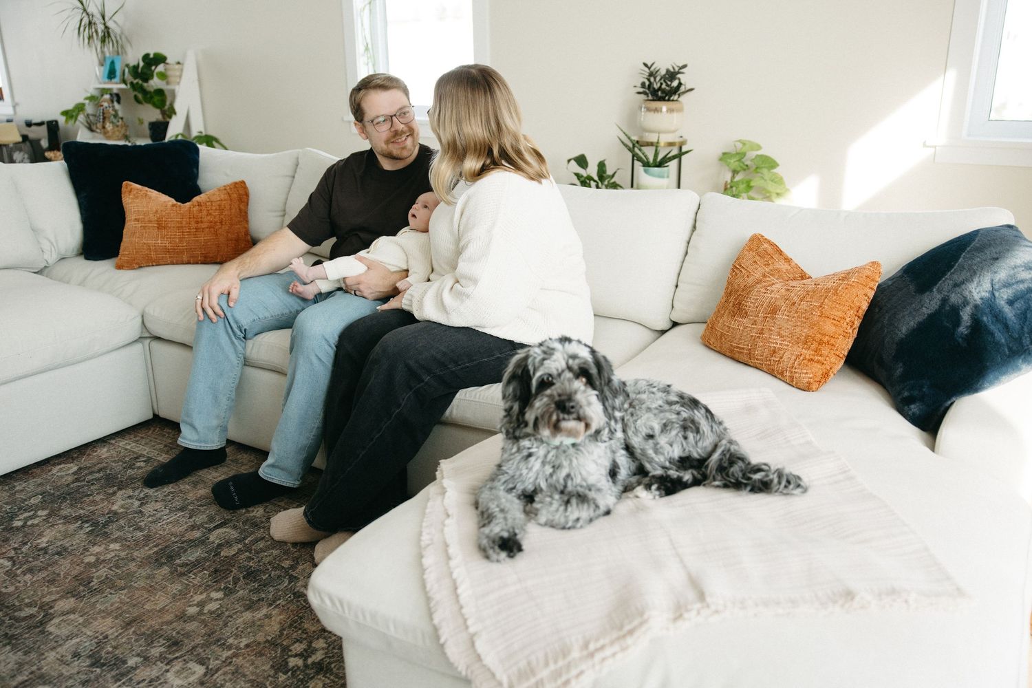 A couple relaxes on a white sectional sofa with orange pillows while their spotted dog lounges beside them.