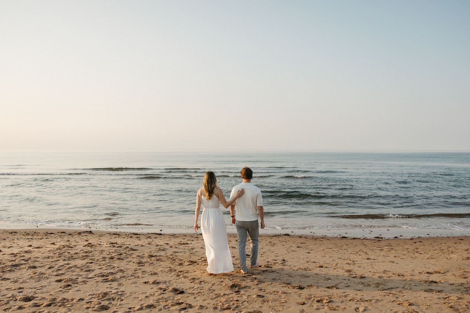 Couple holding hands walking on sandy beach toward calm ocean at sunset in white outfits.