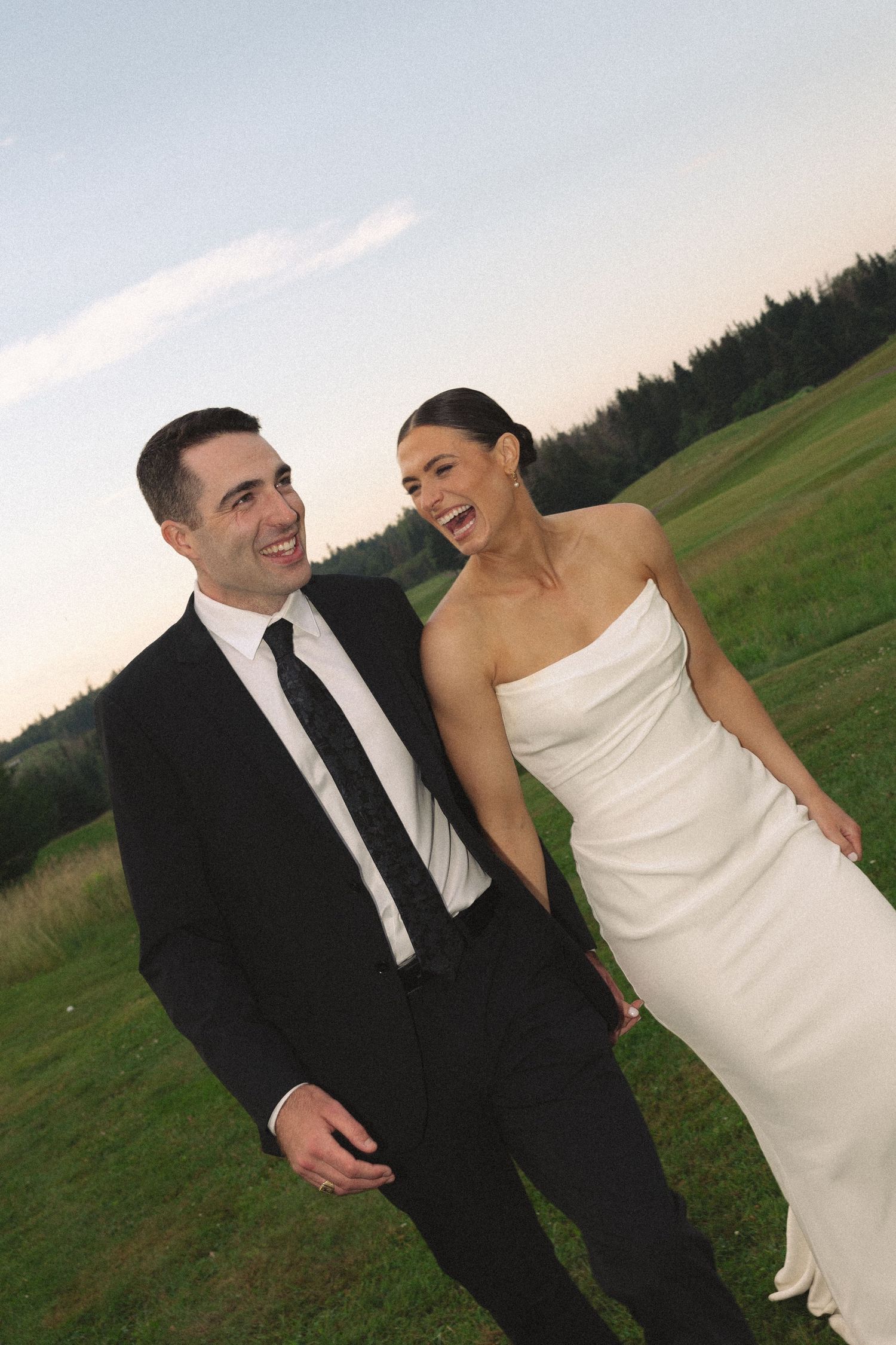 Happy bride and groom laughing together outdoors at dusk, wearing white gown and black suit on green field.