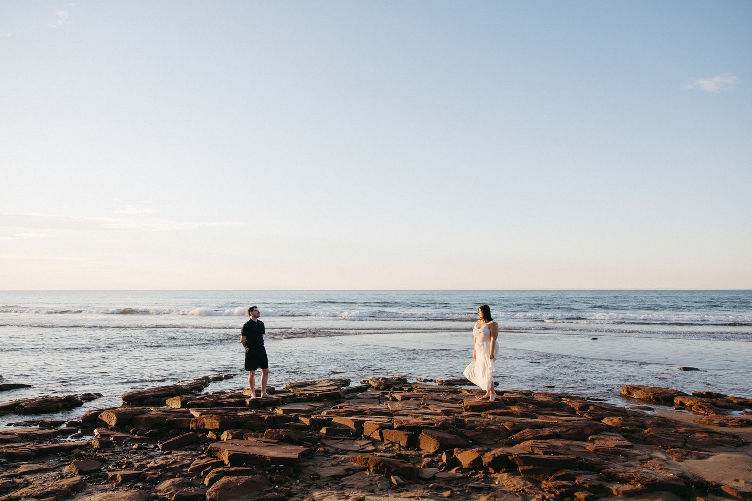 A couple shares a romantic moment on rocky coastal shores with calm ocean and clear blue sky behind.