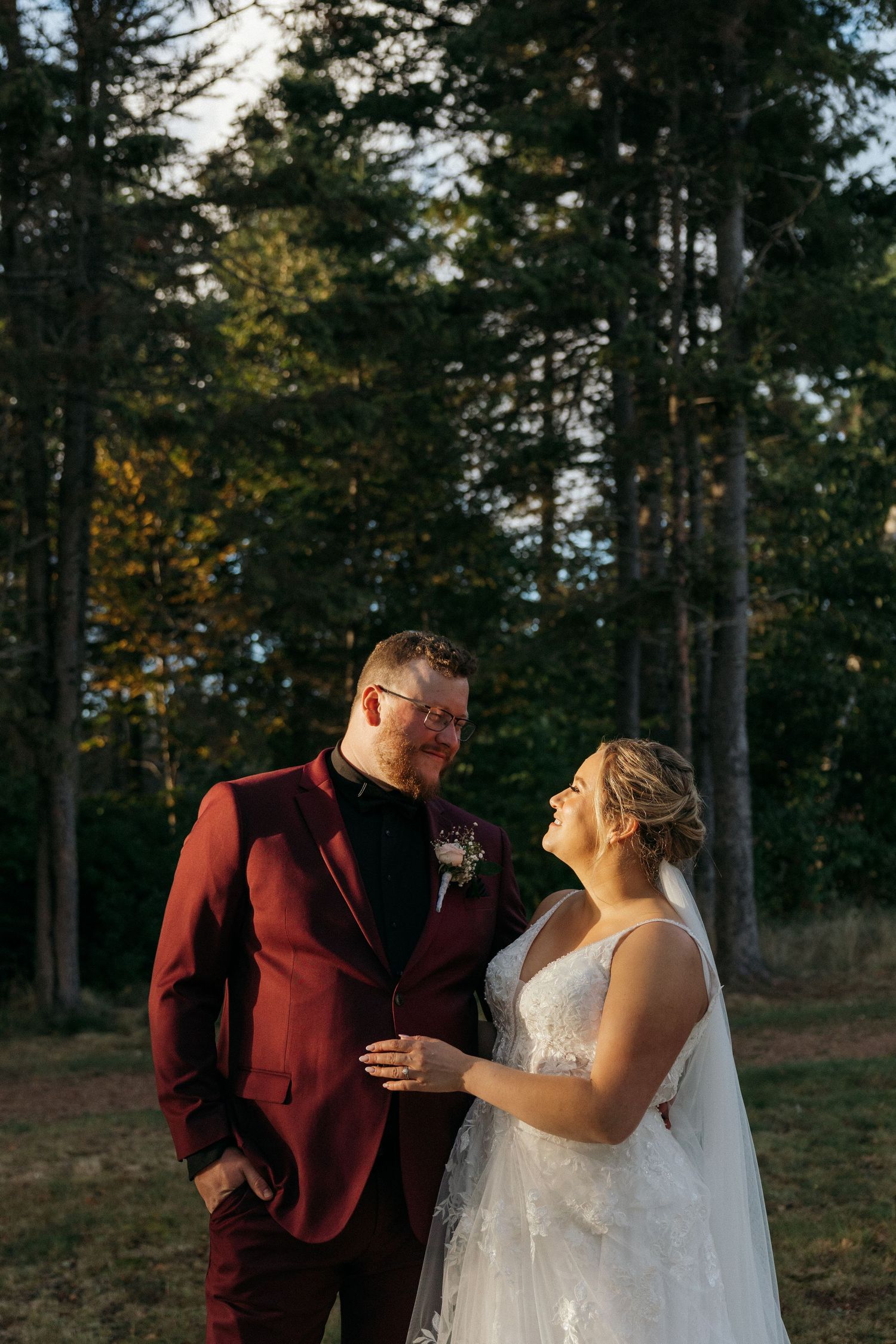 A couple in burgundy and white wedding attire share a joyful moment in a forest setting.