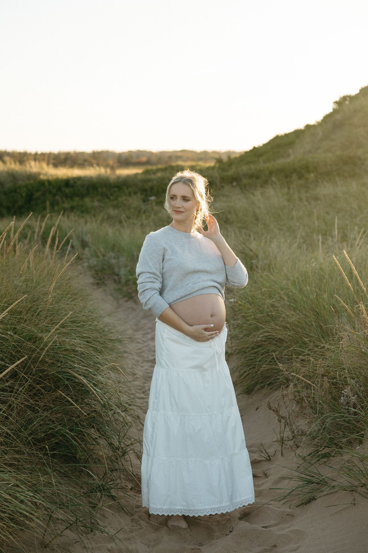 Maternity photo session in sand dunes with soft lighting and flowing white dress against natural coastal backdrop.