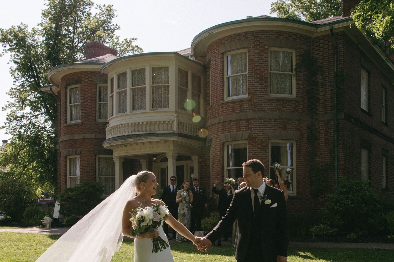 A wedding couple stands together on the grounds of a stately brick mansion with curved architecture.