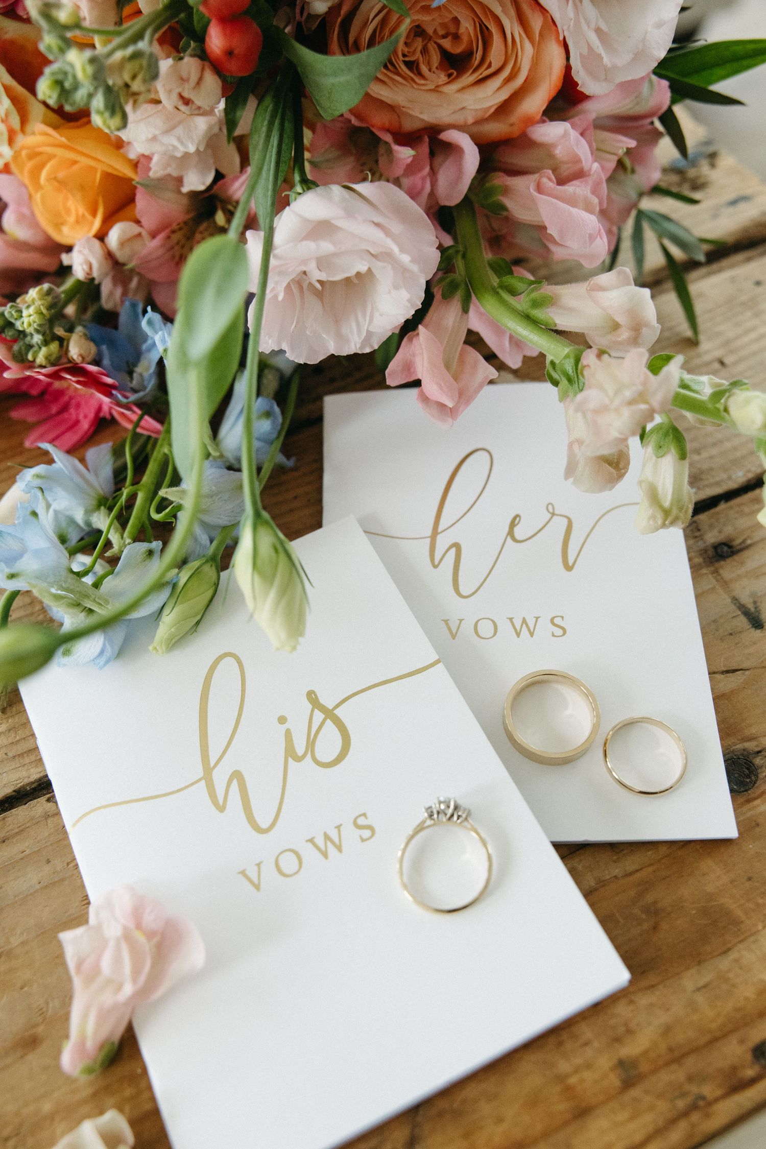 Wedding vow books with gold lettering surrounded by pink and peach florals and wedding rings.