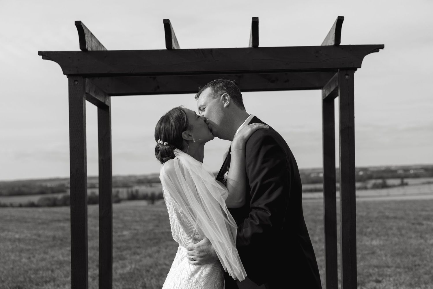 A romantic outdoor wedding ceremony with a couple embracing under a wooden arch structure.
