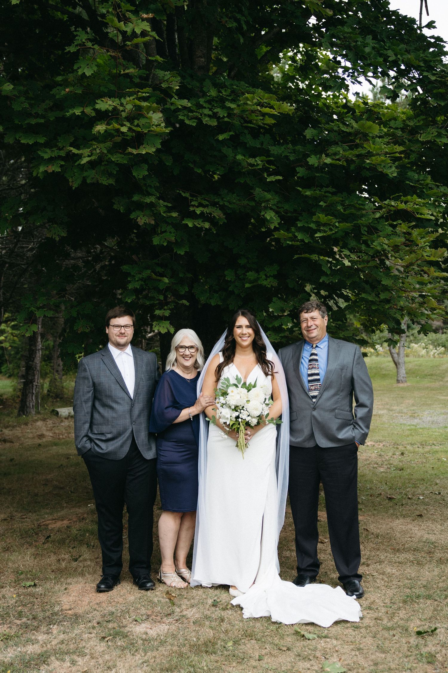Wedding group photo with guests in formal attire gathered outdoors under tree canopy.