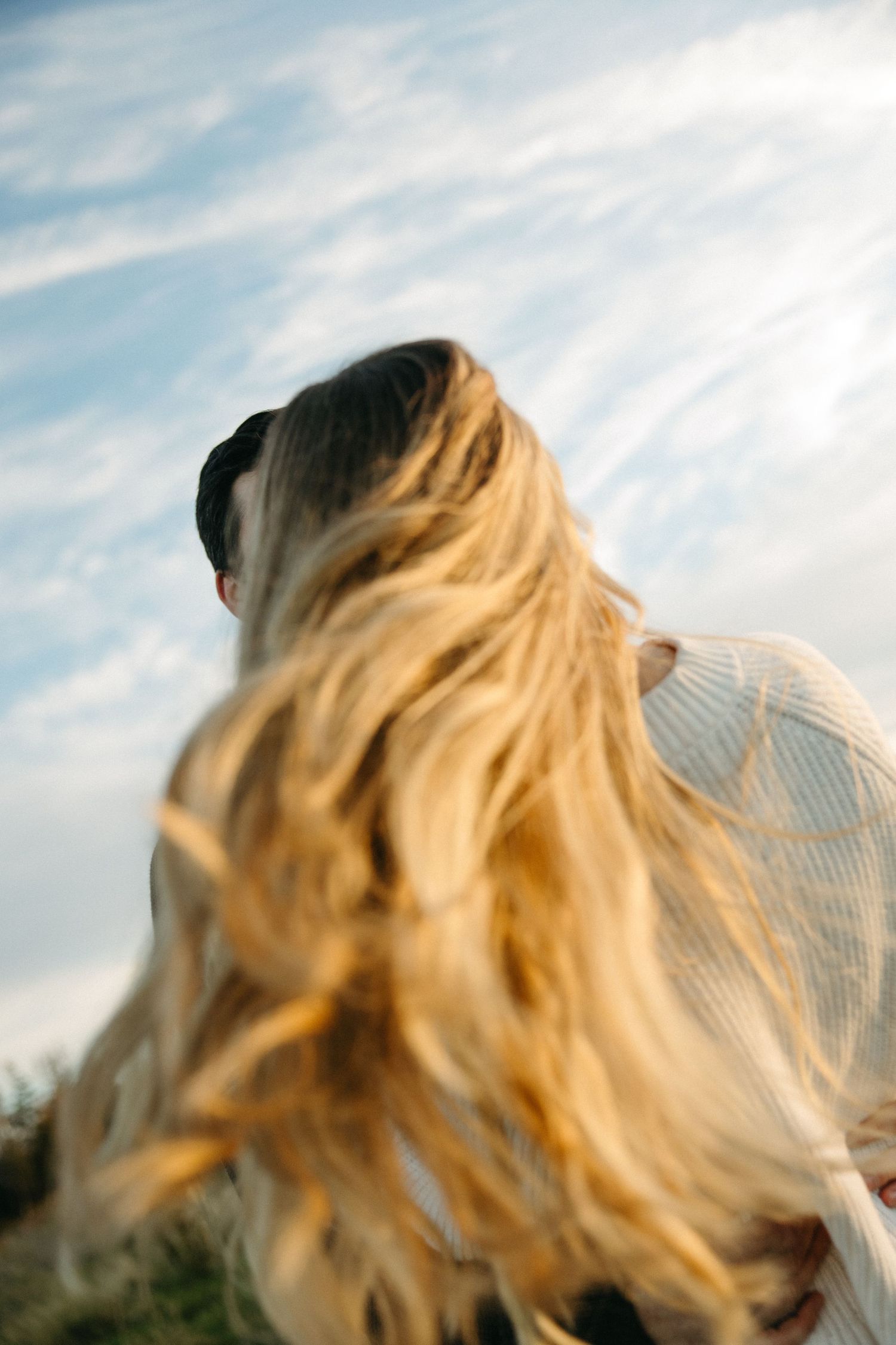 Woman with long wind-blown blonde hair embracing a man outdoors under a blue sky with soft wispy clouds.