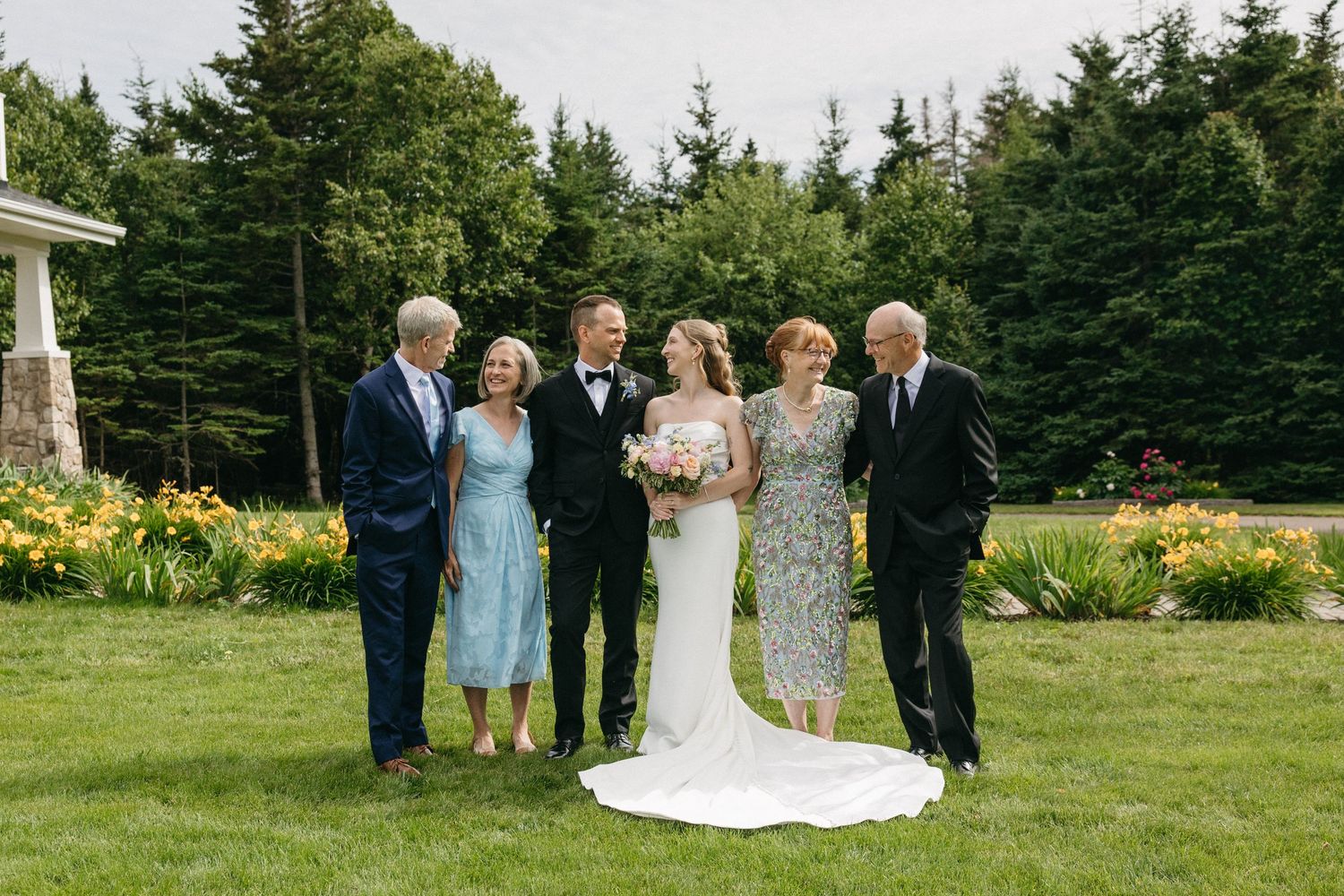Wedding party group photo with bride and groom surrounded by family in a lush garden with tall evergreen trees.