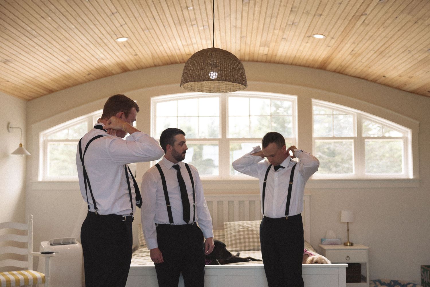 Three groomsmen in white shirts and black suspenders getting ready together in a cozy wooden room.