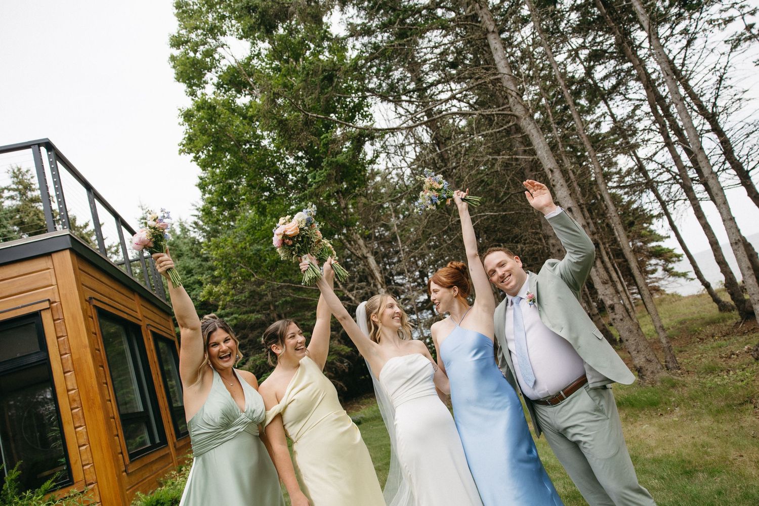 Wedding party celebrating outdoors with raised arms, bride in white gown with bridesmaids in pastel dresses near trees.