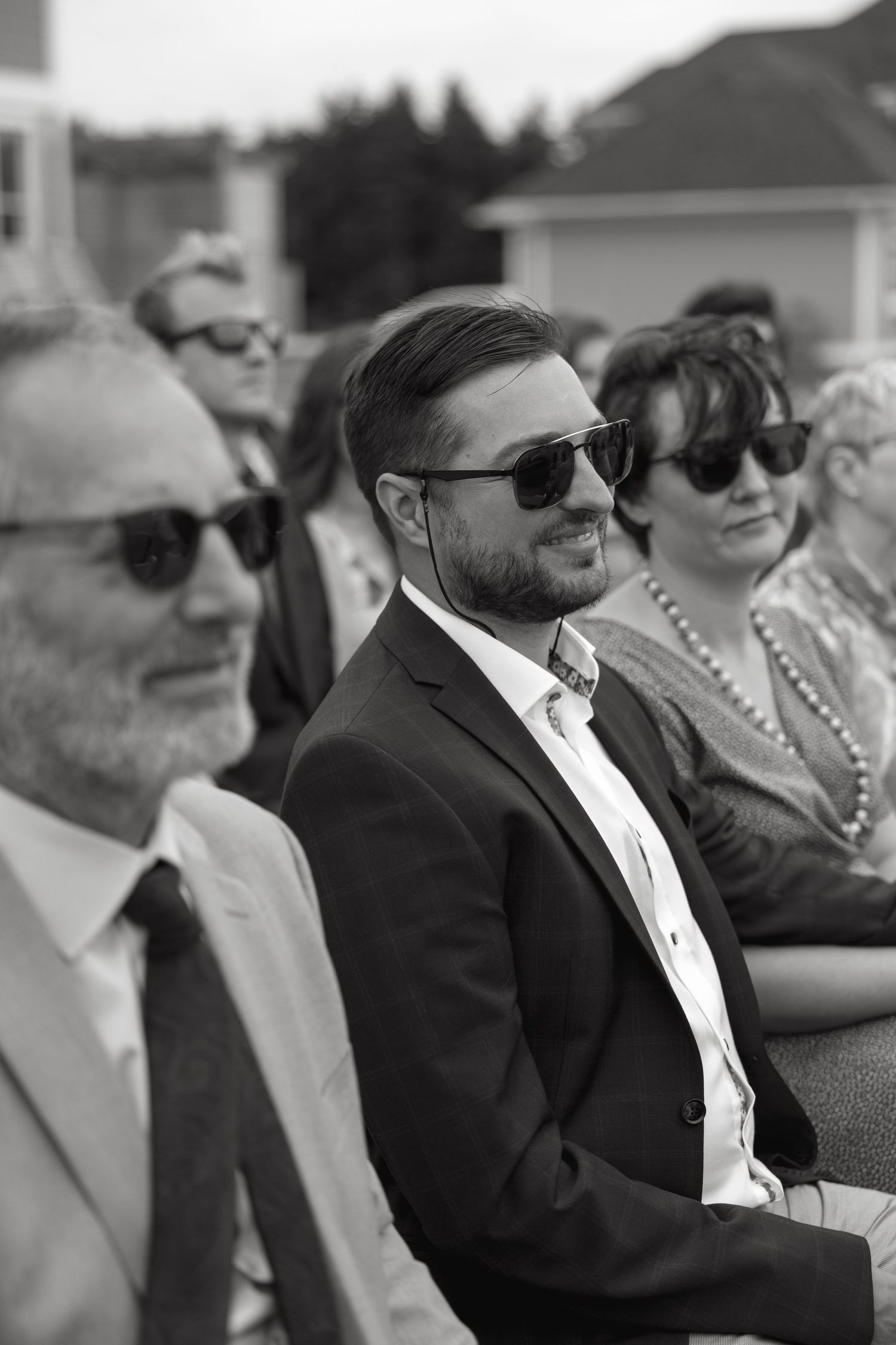 Black and white photo of smiling wedding guests wearing sunglasses seated at outdoor ceremony.