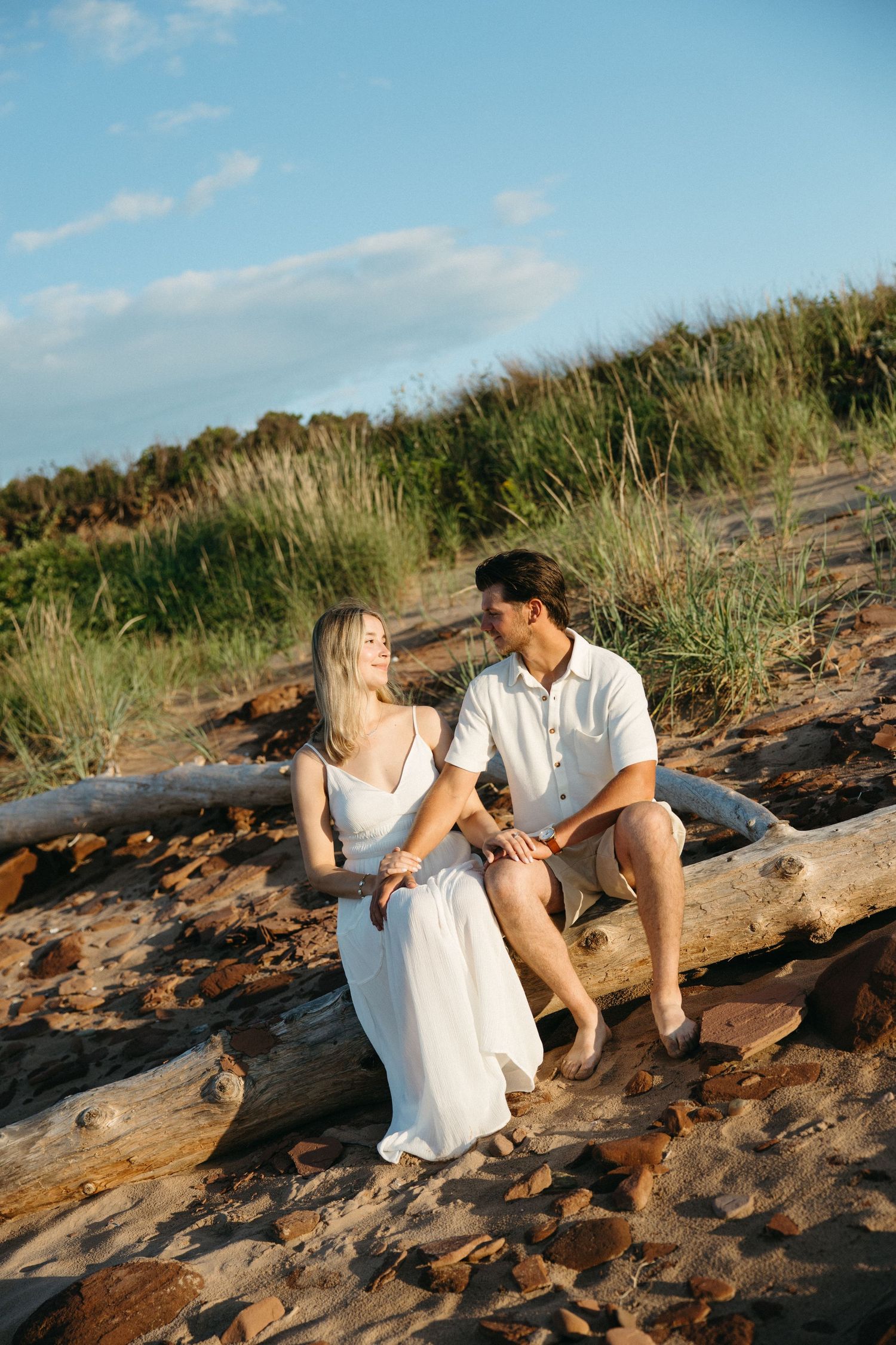 Couple sitting on driftwood log on sandy beach, woman in white dress, man in white shirt, golden hour light.