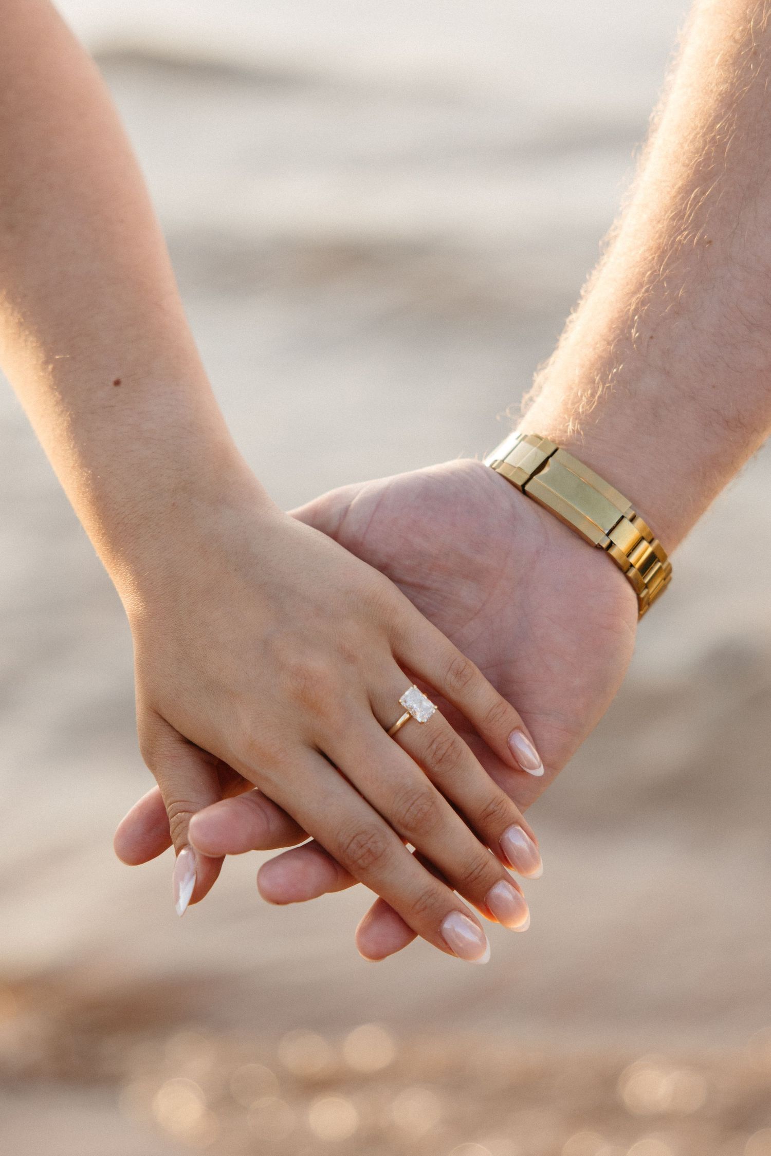 Close-up of engaged couple holding hands on beach, woman wearing emerald-cut diamond ring, man wearing gold watch.