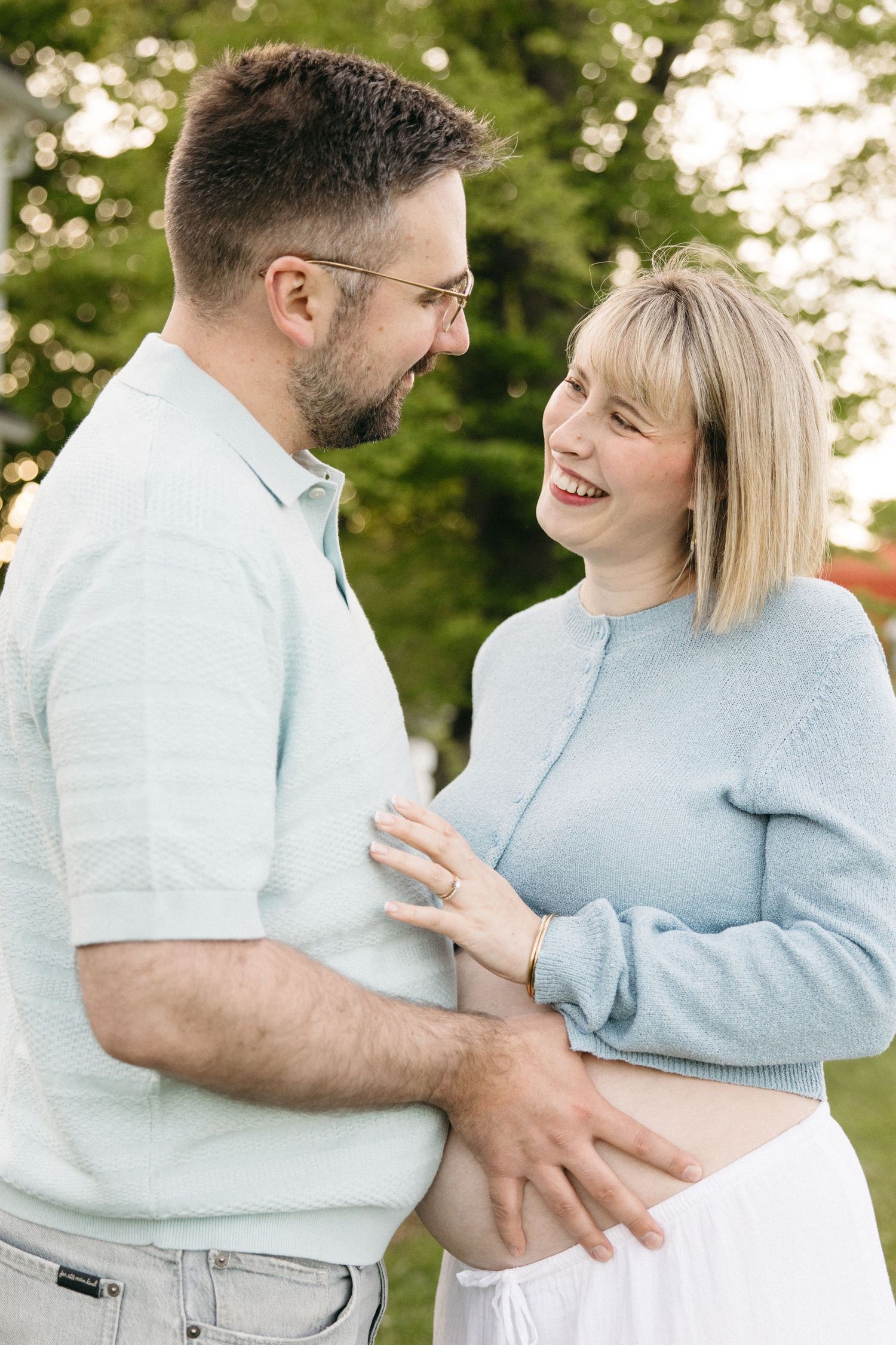 Smiling pregnant couple in matching light blue outfits holding baby bump outdoors with green trees in background.