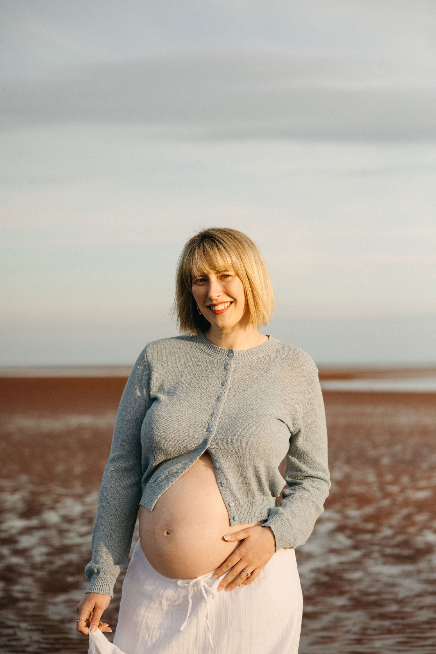 Smiling pregnant woman in gray cardigan holding baby bump on a beach at golden hour.