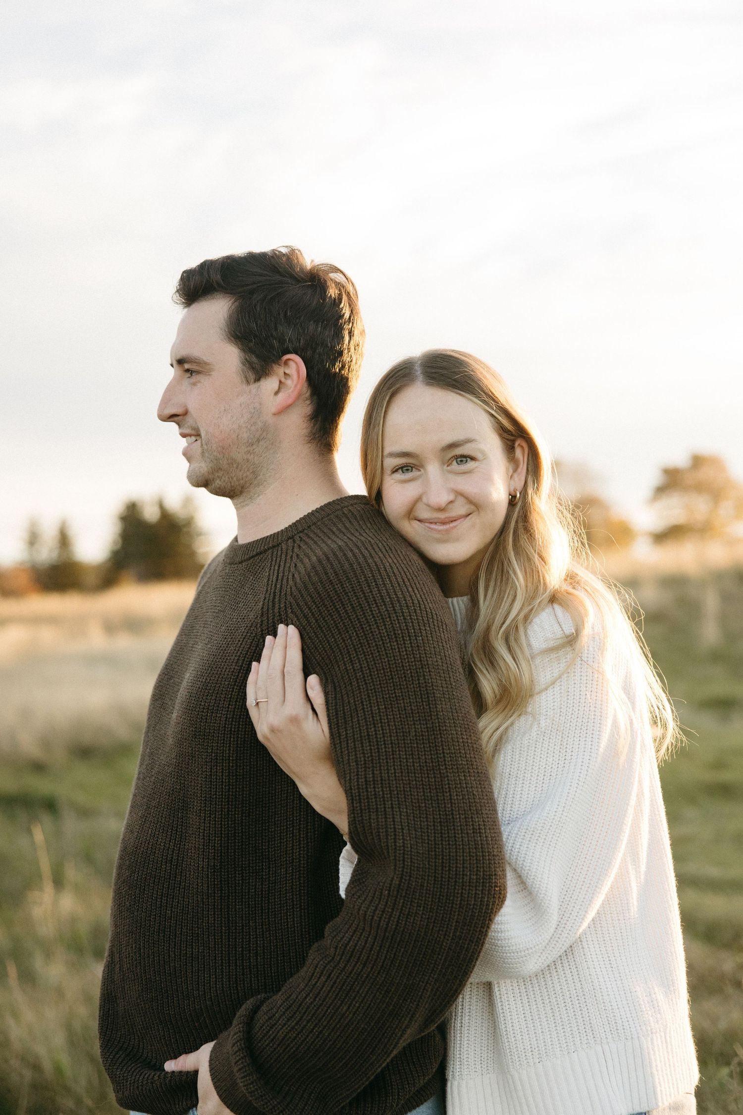 Man in a brown knit sweater looks aside as a smiling woman in a cream sweater hugs him from behind in a golden field at sunset.
