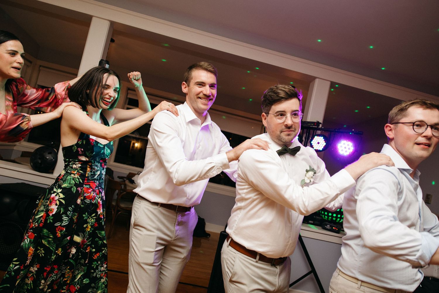 Wedding guests form a dance line on the reception dance floor under colorful party lights.