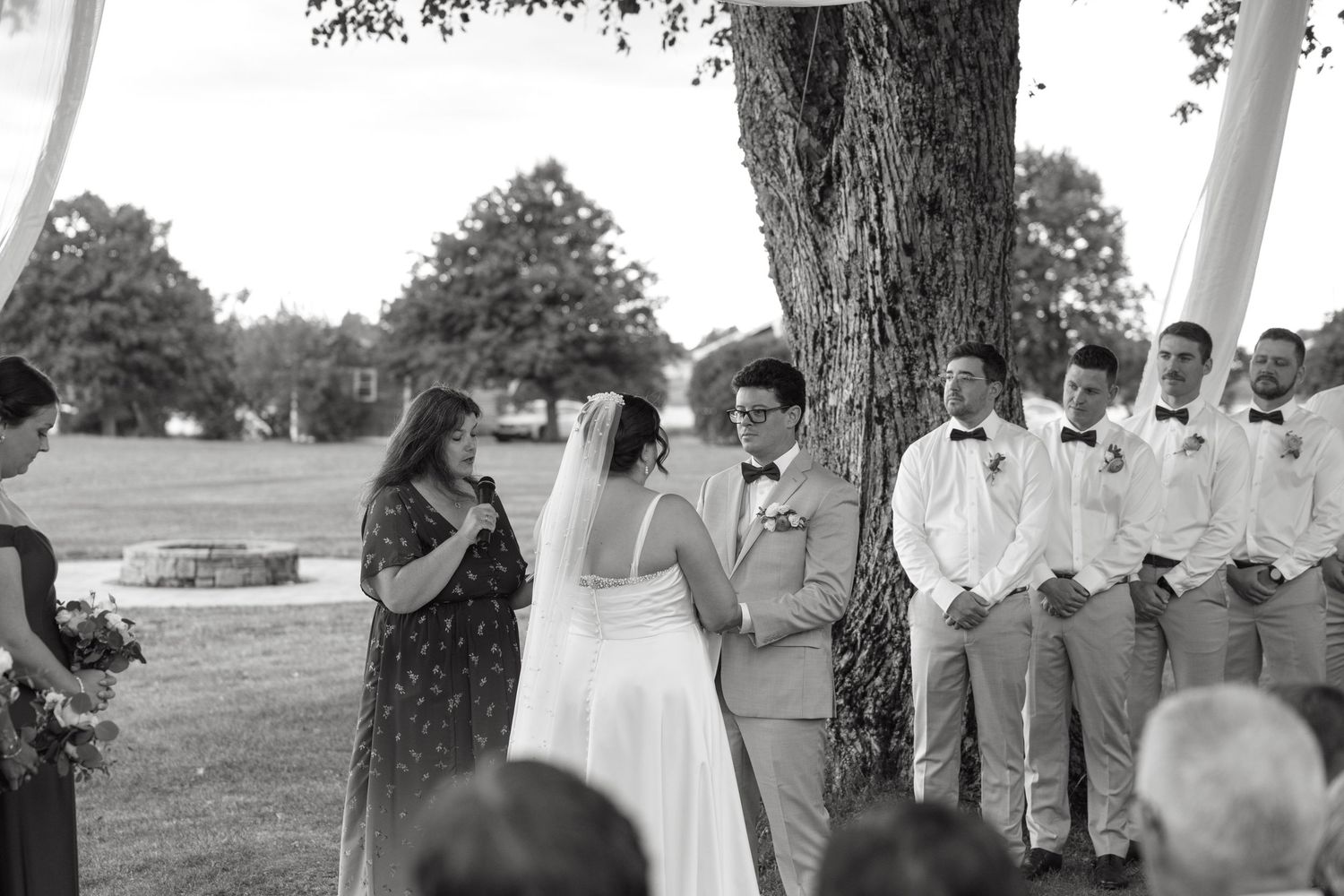 Bride and groom exchange vows outdoors under a tree with groomsmen standing nearby during wedding ceremony.