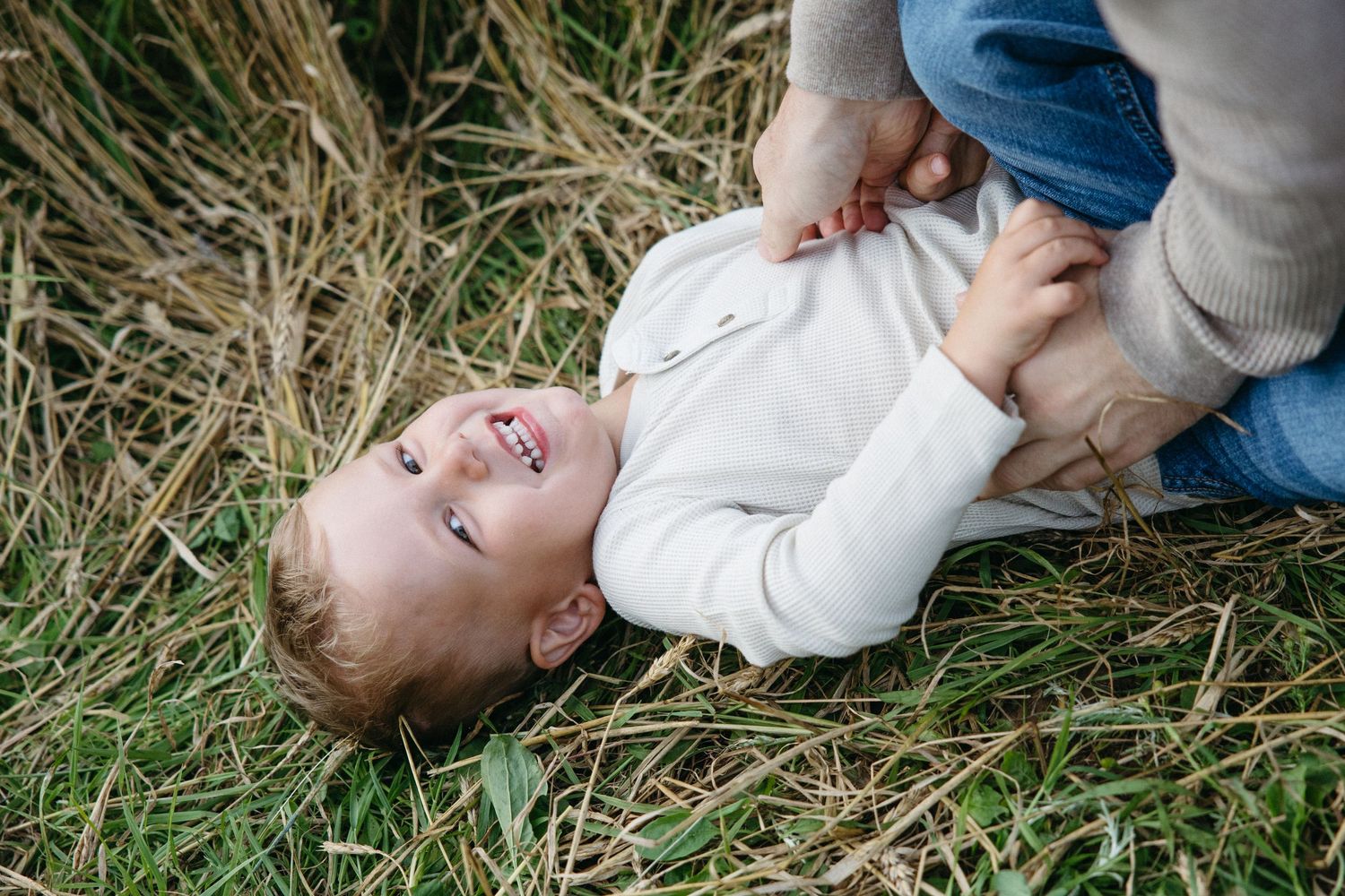 A laughing toddler lies on grass while an adult playfully tickles them outdoors.