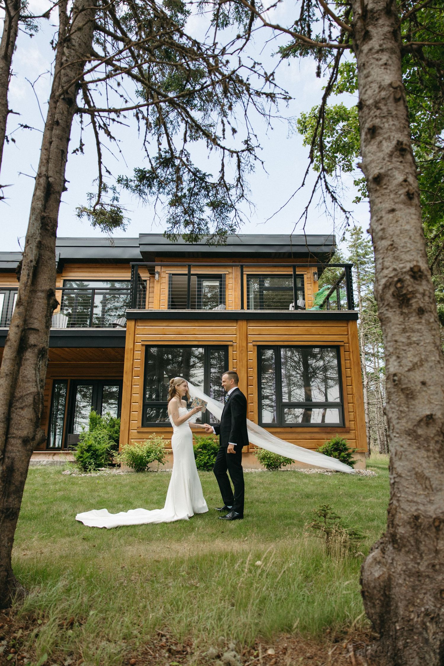 A bride and groom share a romantic moment in front of a modern wooden house surrounded by trees.