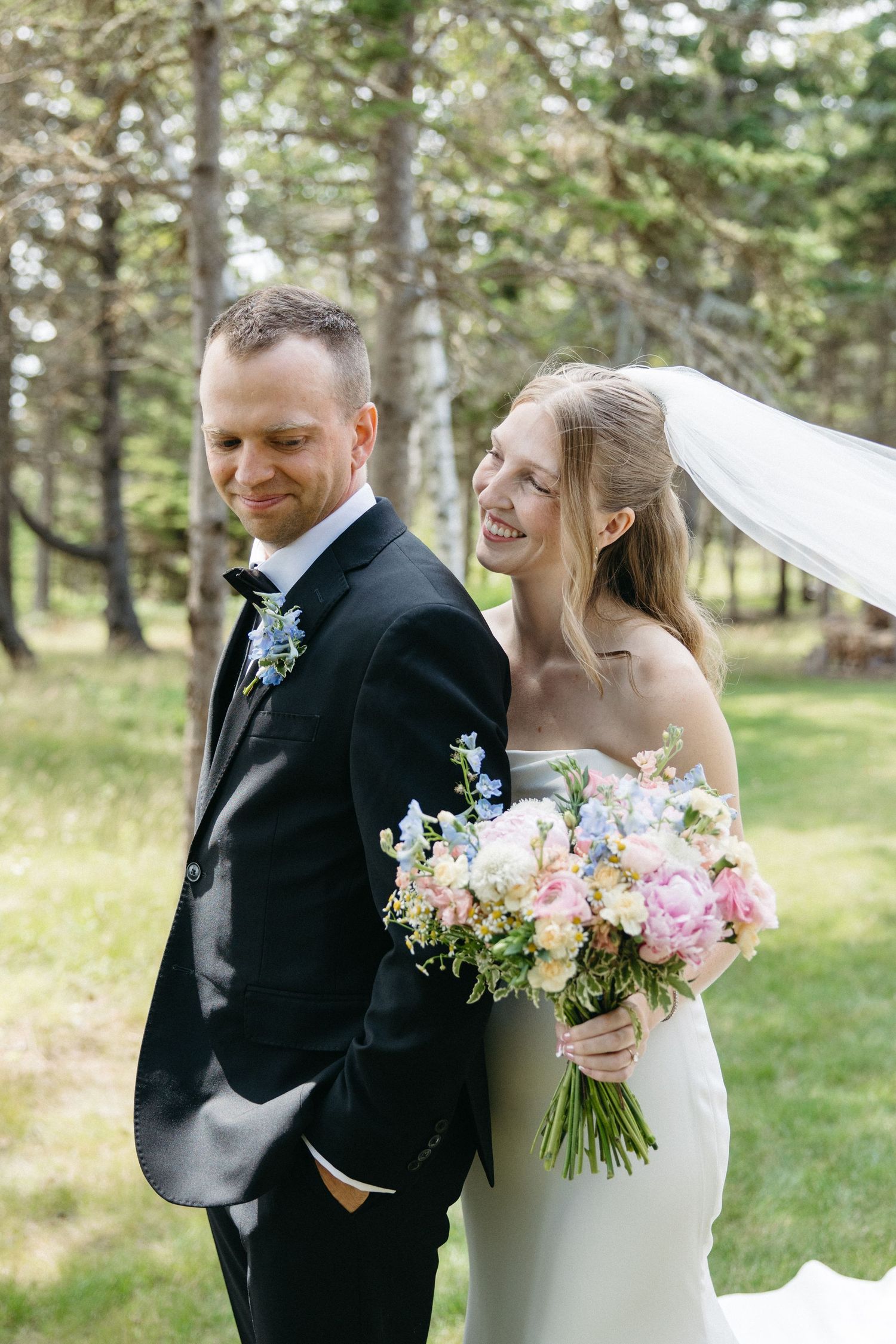 Smiling bride in strapless gown holds pastel bouquet standing back-to-back with groom in black tuxedo.