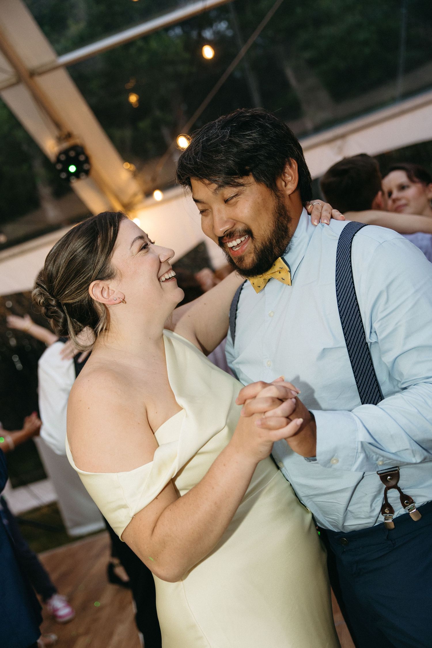 Bride in ivory gown and groom in light blue shirt with yellow bow tie share a joyful first dance under string lights.