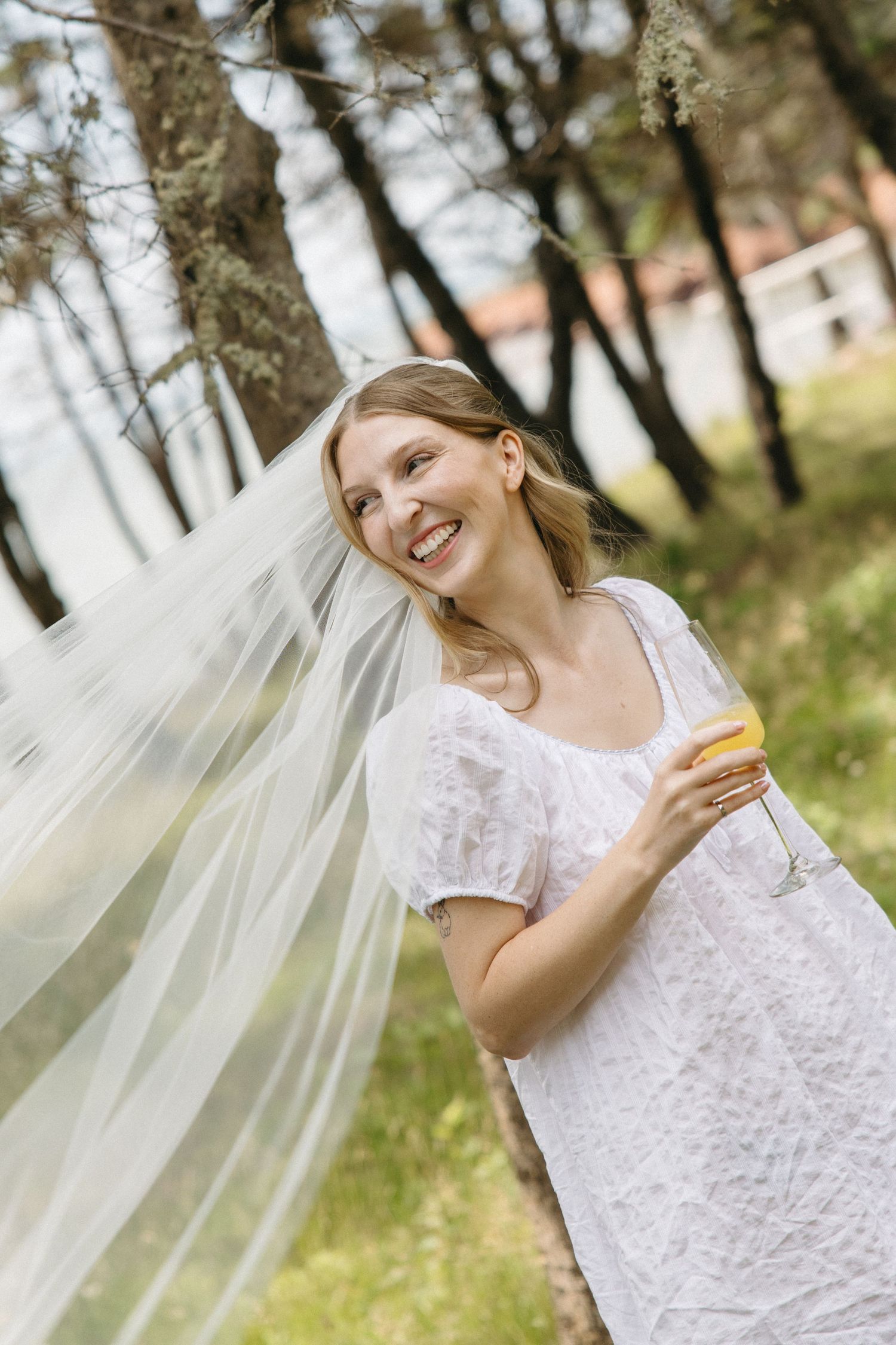 Smiling bride in white dress holding champagne glass outdoors with veil flowing in the breeze among trees.